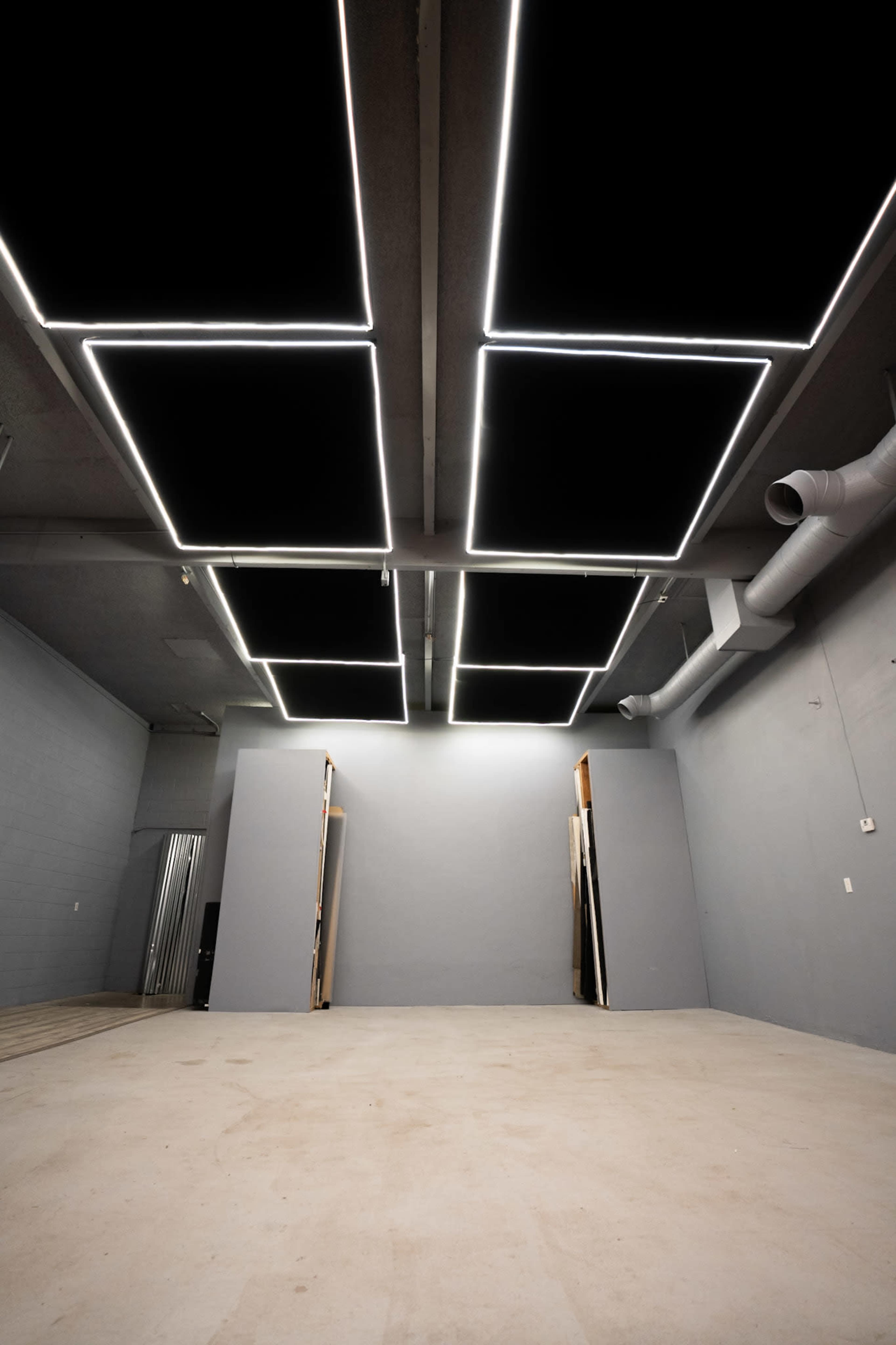 The image shows an empty room with a gray wall, bare floor, and illuminated rectangular light fixtures on the ceiling.