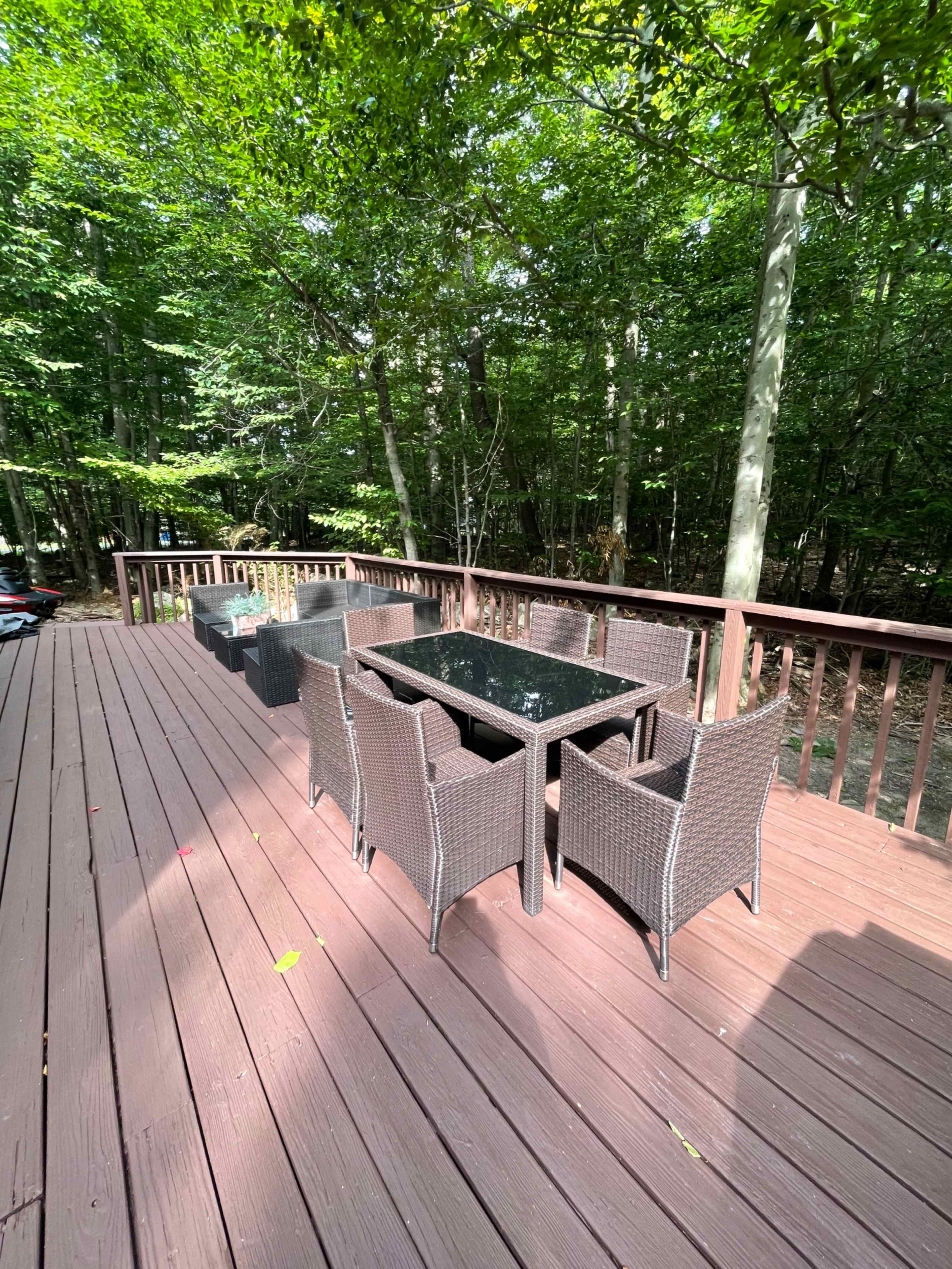 A wooden deck surrounded by trees features a glass-top dining table with eight wicker chairs.