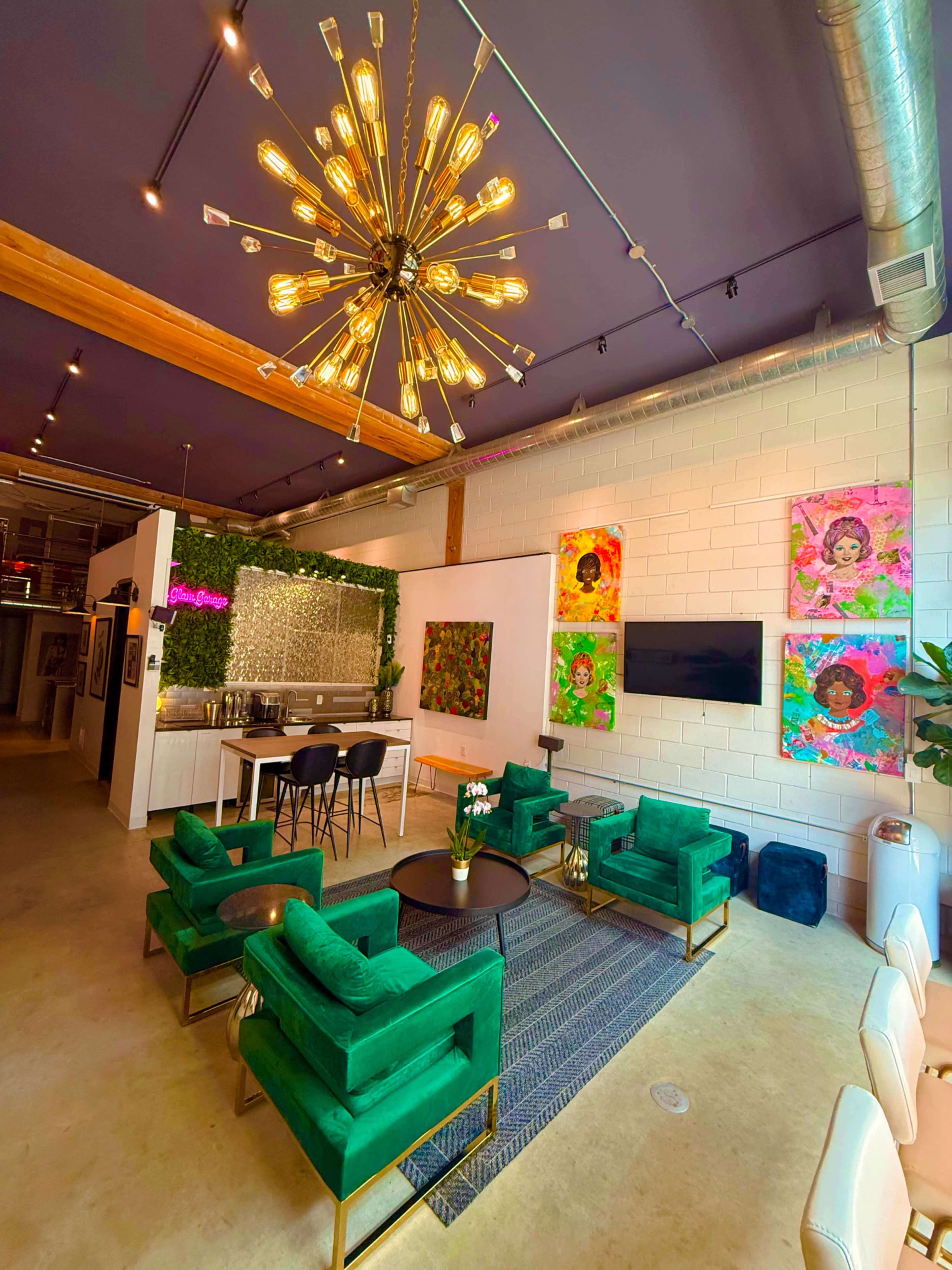 The image shows a brightly lit modern lounge area featuring green velvet chairs, a contemporary chandelier, and colorful artworks on the walls.