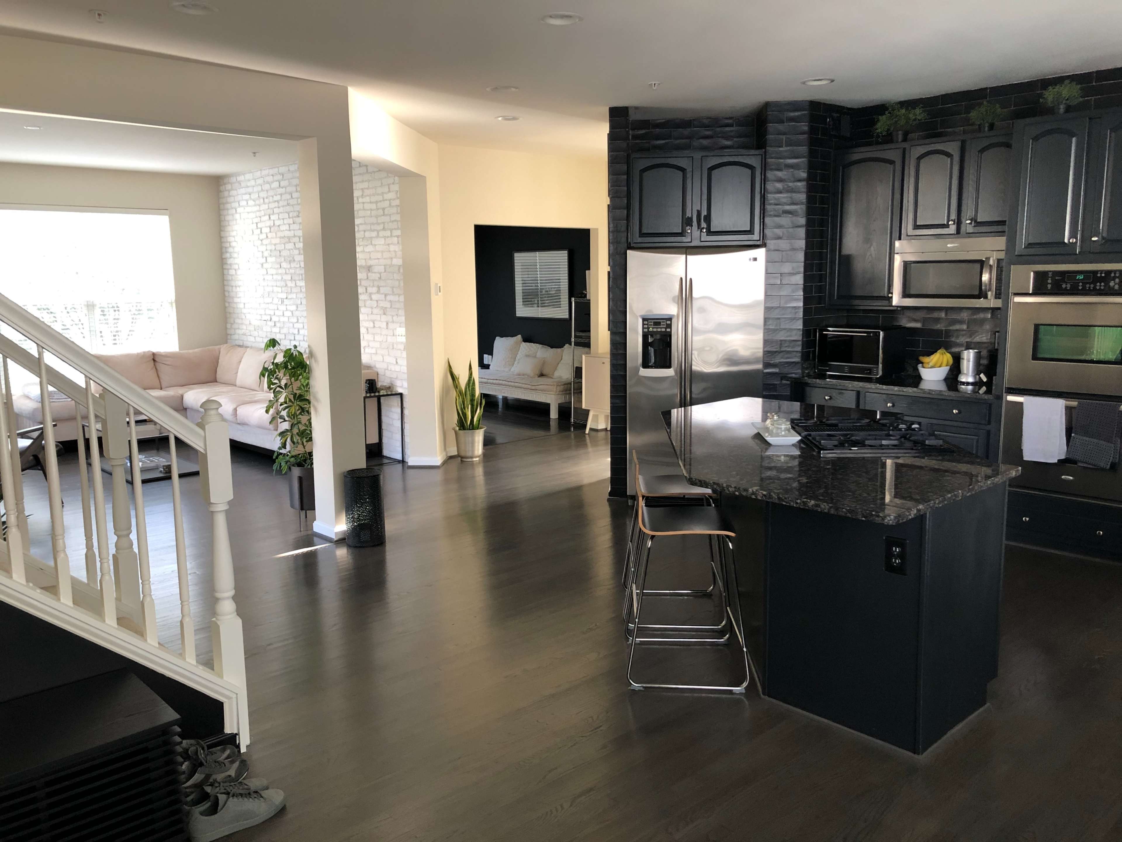 A modern kitchen with dark cabinetry, a granite countertop, and a view into a light-filled living area featuring a sofa and plants.