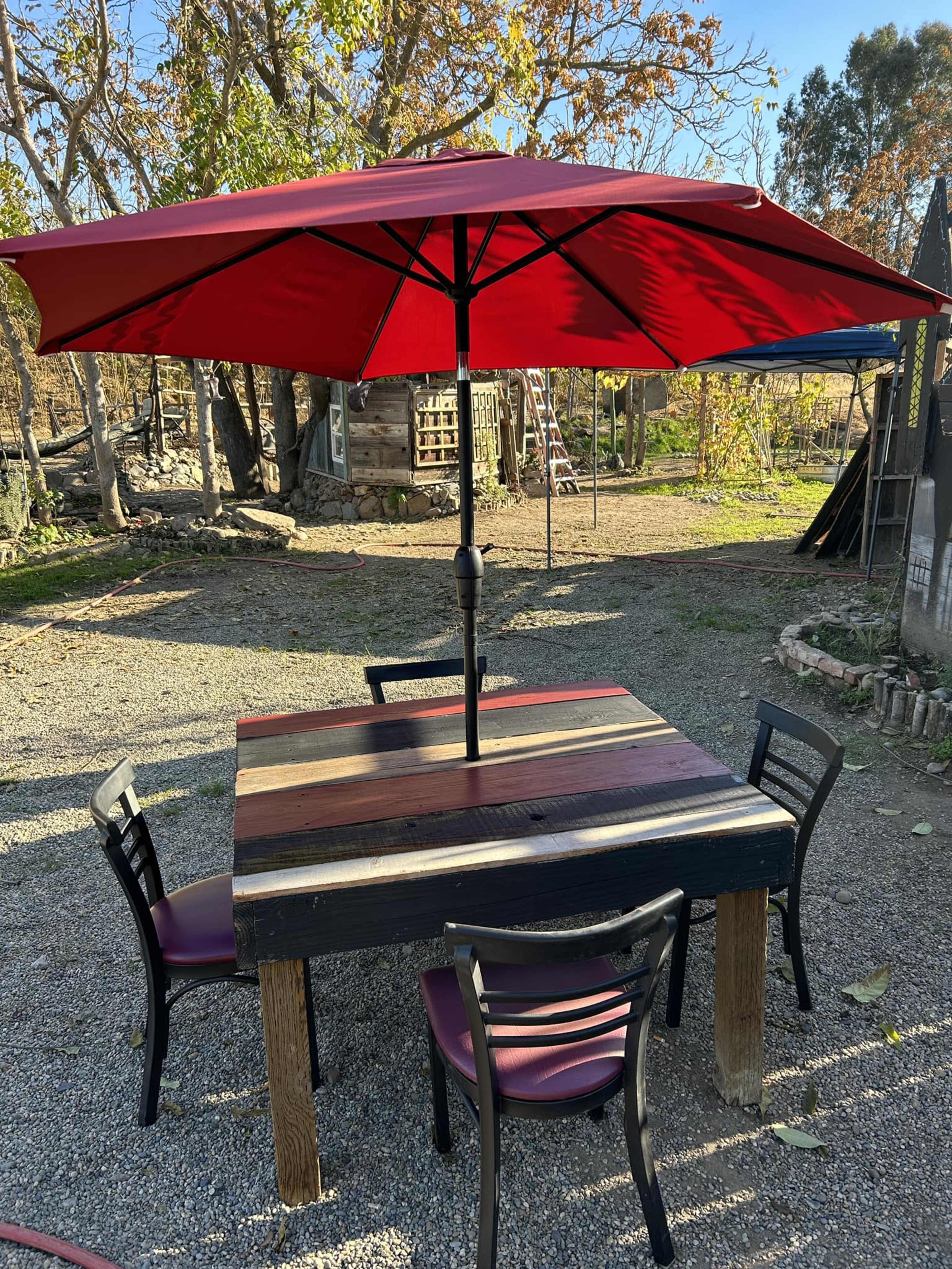 A wooden table with a red umbrella is surrounded by four black chairs on a gravel surface in an outdoor setting.
