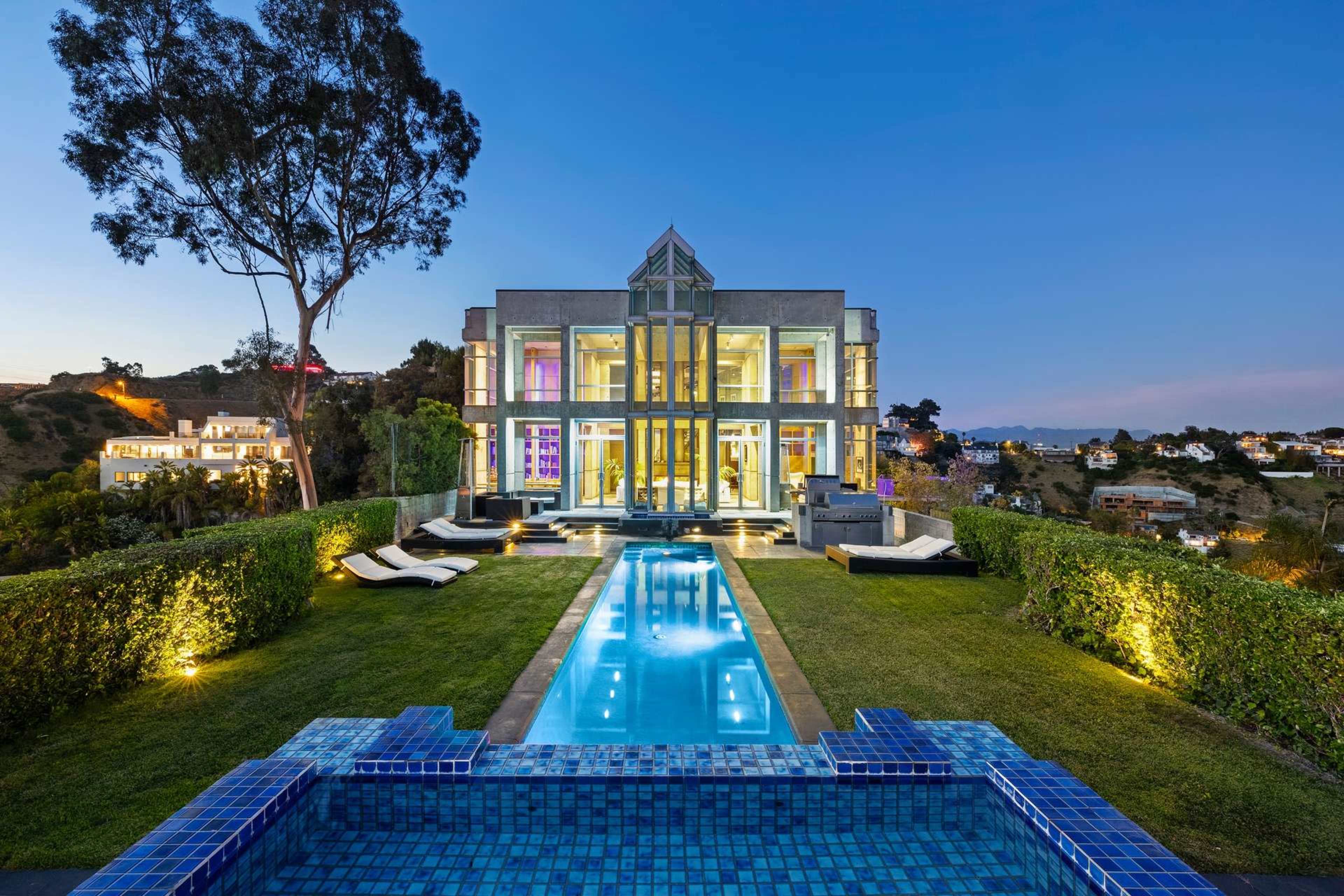 A modern mansion with large glass windows overlooks a swimming pool and manicured lawn under a twilight sky.