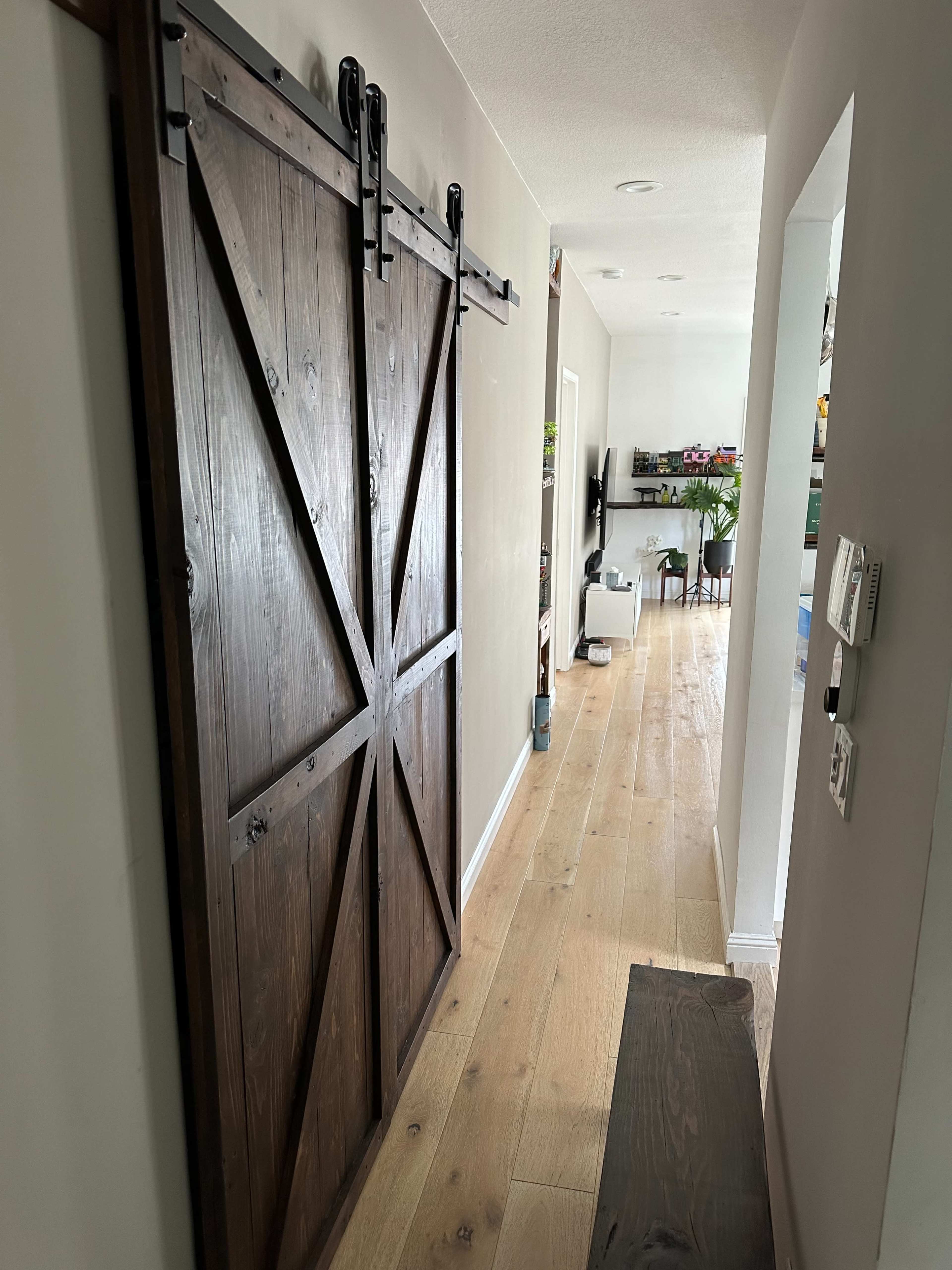 A hallway featuring a wooden barn door on the left, with light-colored flooring and furniture visible in the distance.