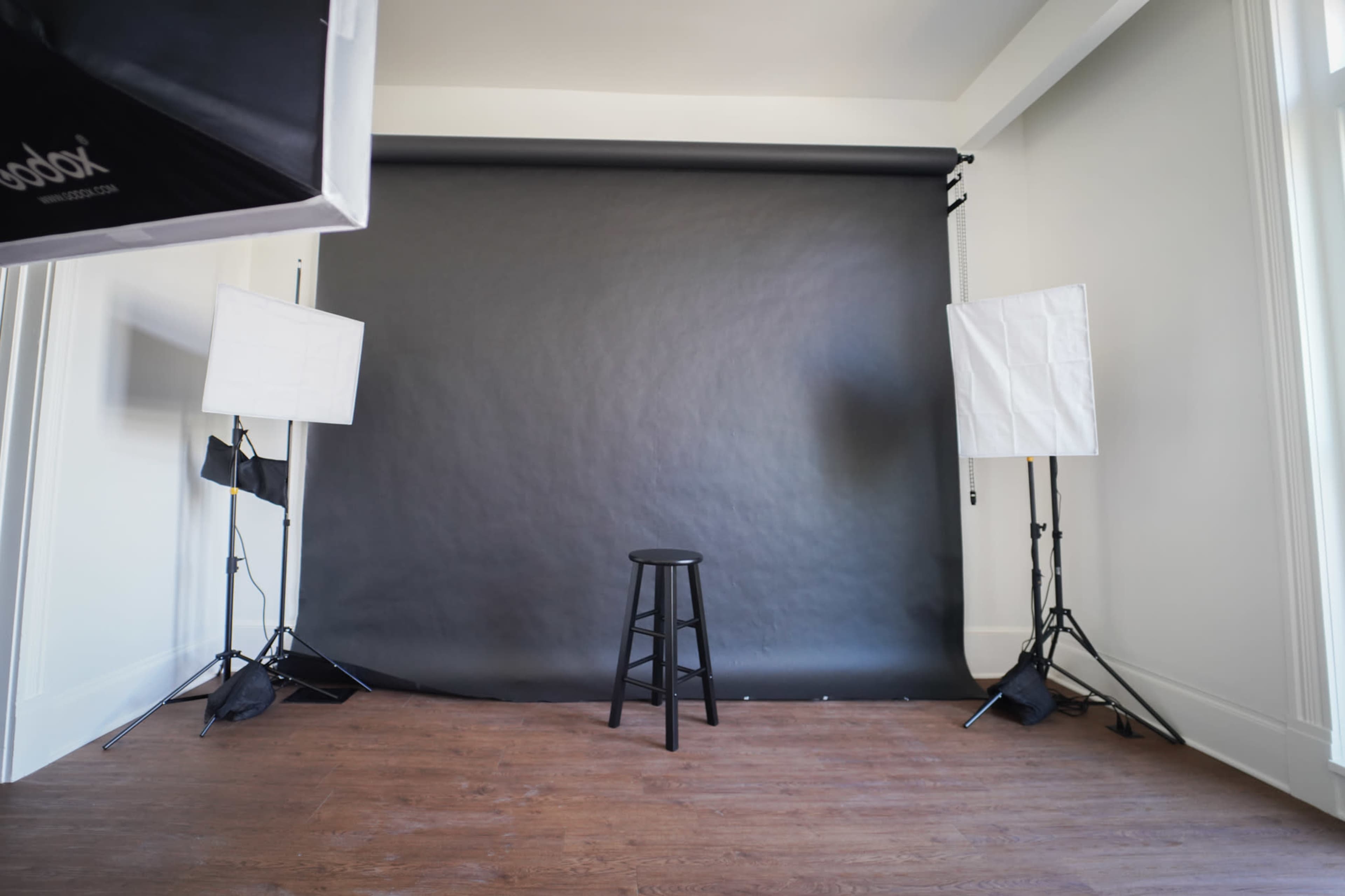 The image shows a photography studio with a black backdrop, an empty stool in the center, and two softbox lights positioned on either side.