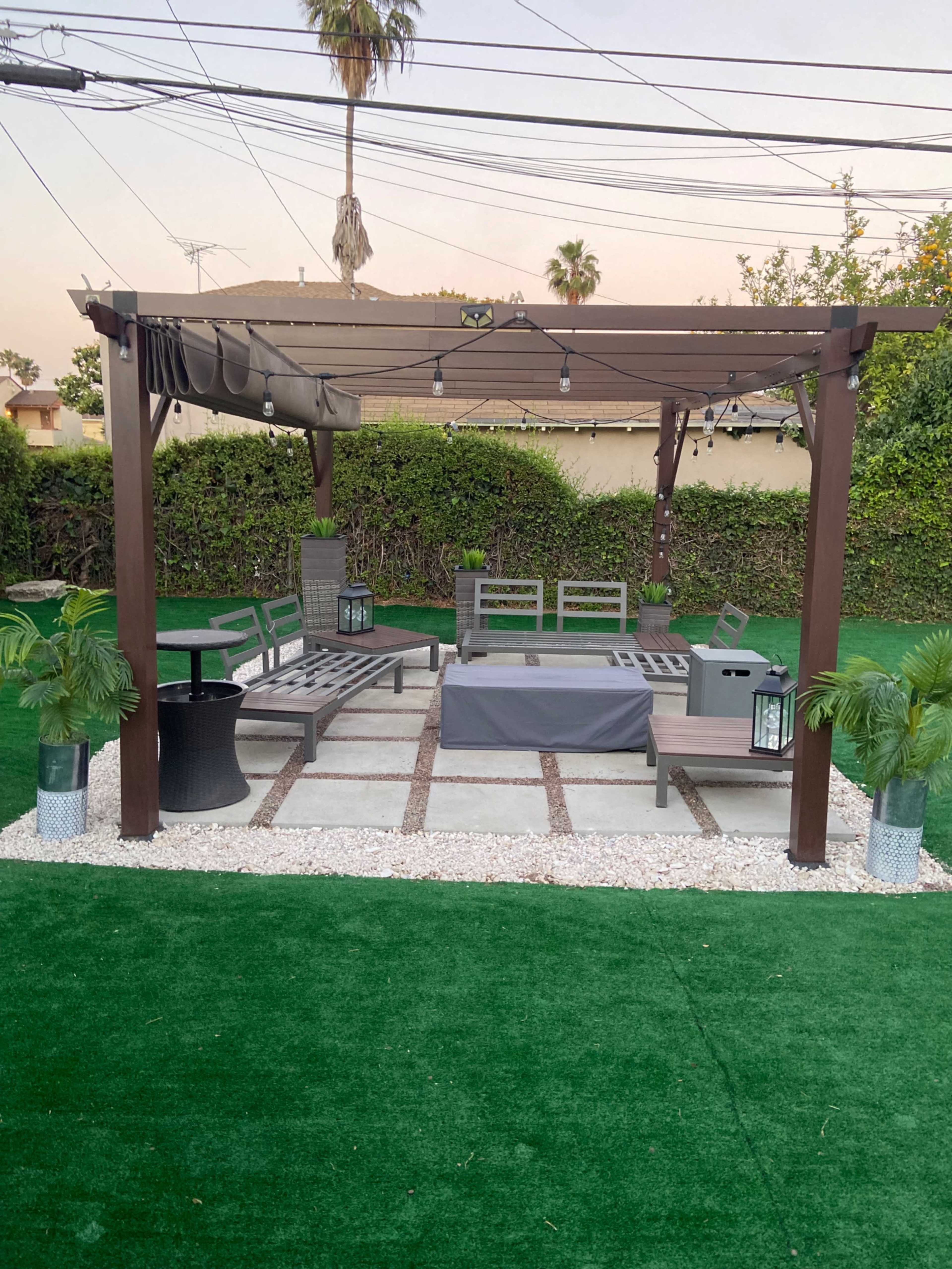 The image shows a gazebo with seating and a fire pit area, surrounded by artificial grass and potted plants.