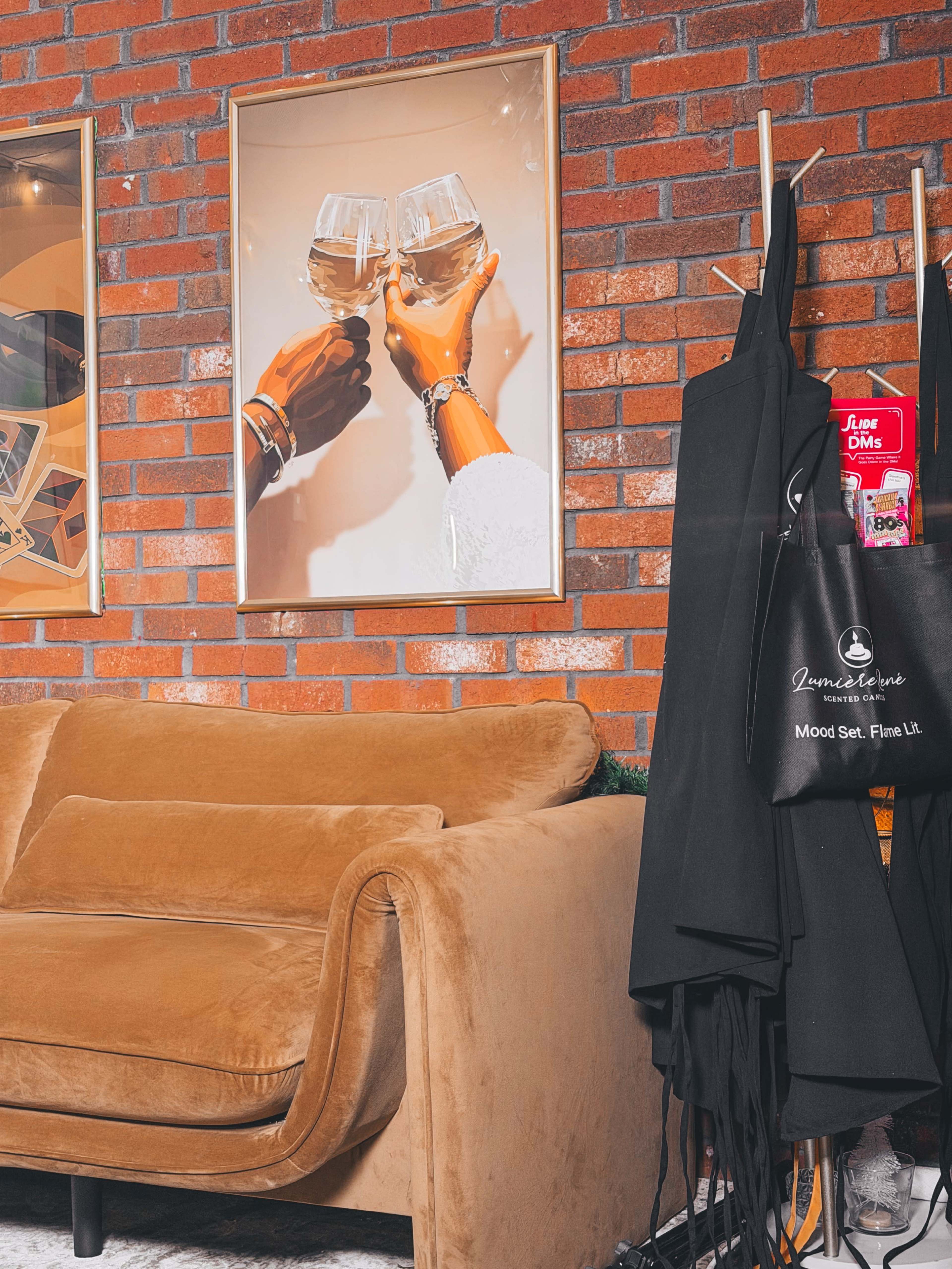 The image shows a cozy seating area featuring a brown couch, a framed artwork of clinking glasses, and a collection of aprons hanging on a brick wall.