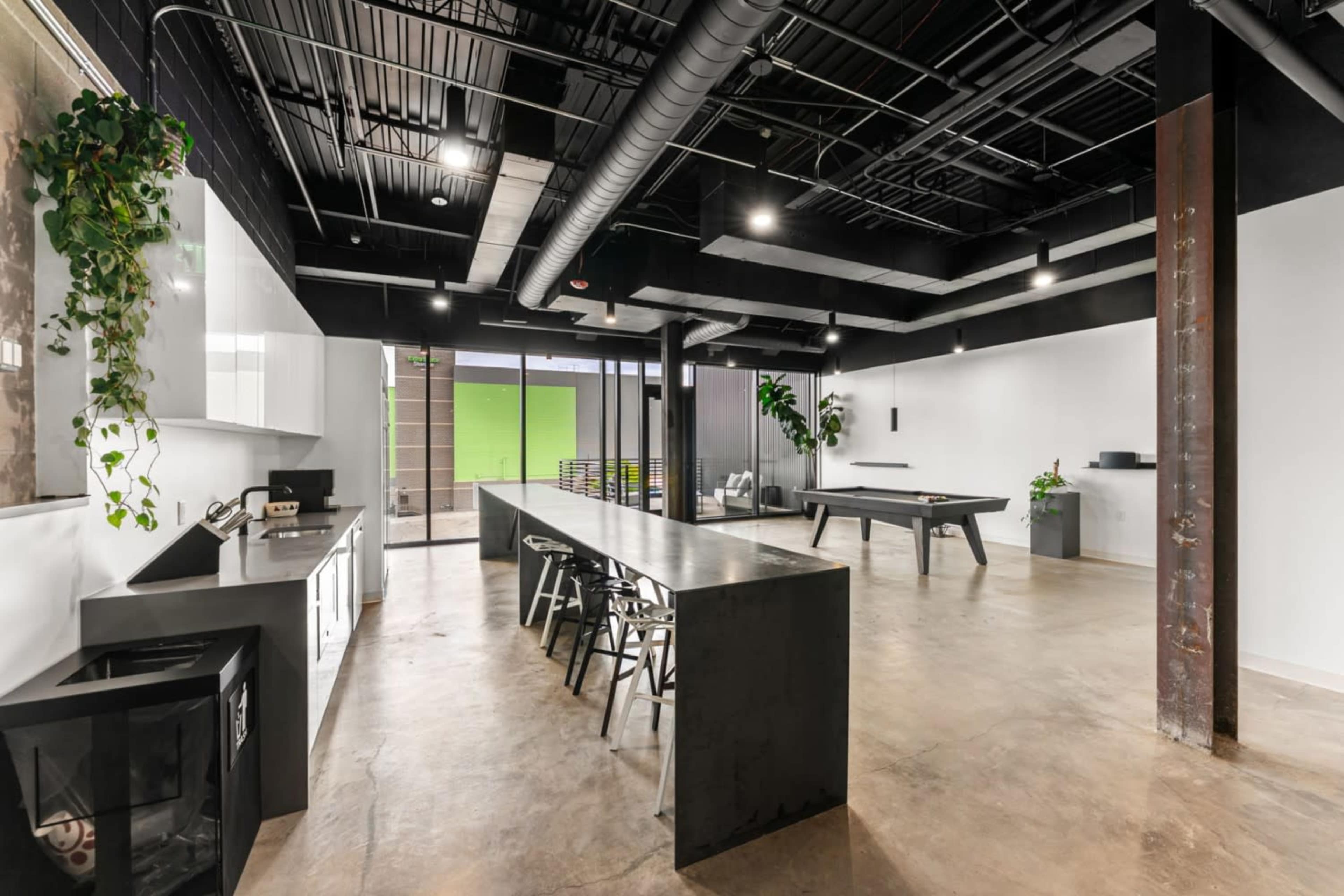The image shows a modern office space featuring a long black table, bar stools, a kitchenette, and a pool table under industrial-style ceilings.