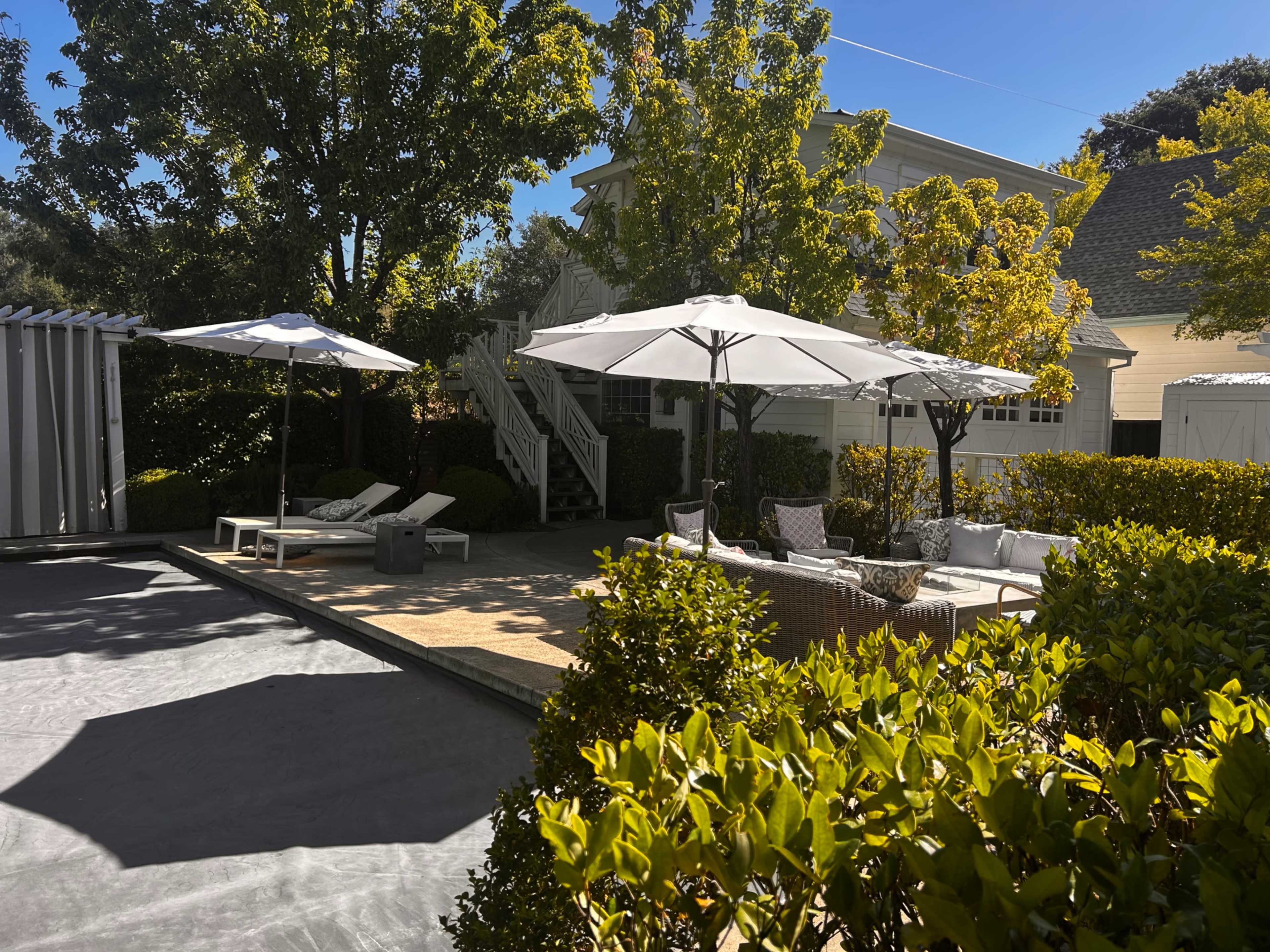 The image shows a landscaped outdoor area featuring umbrellas, lounge chairs, and a seating arrangement surrounded by greenery.