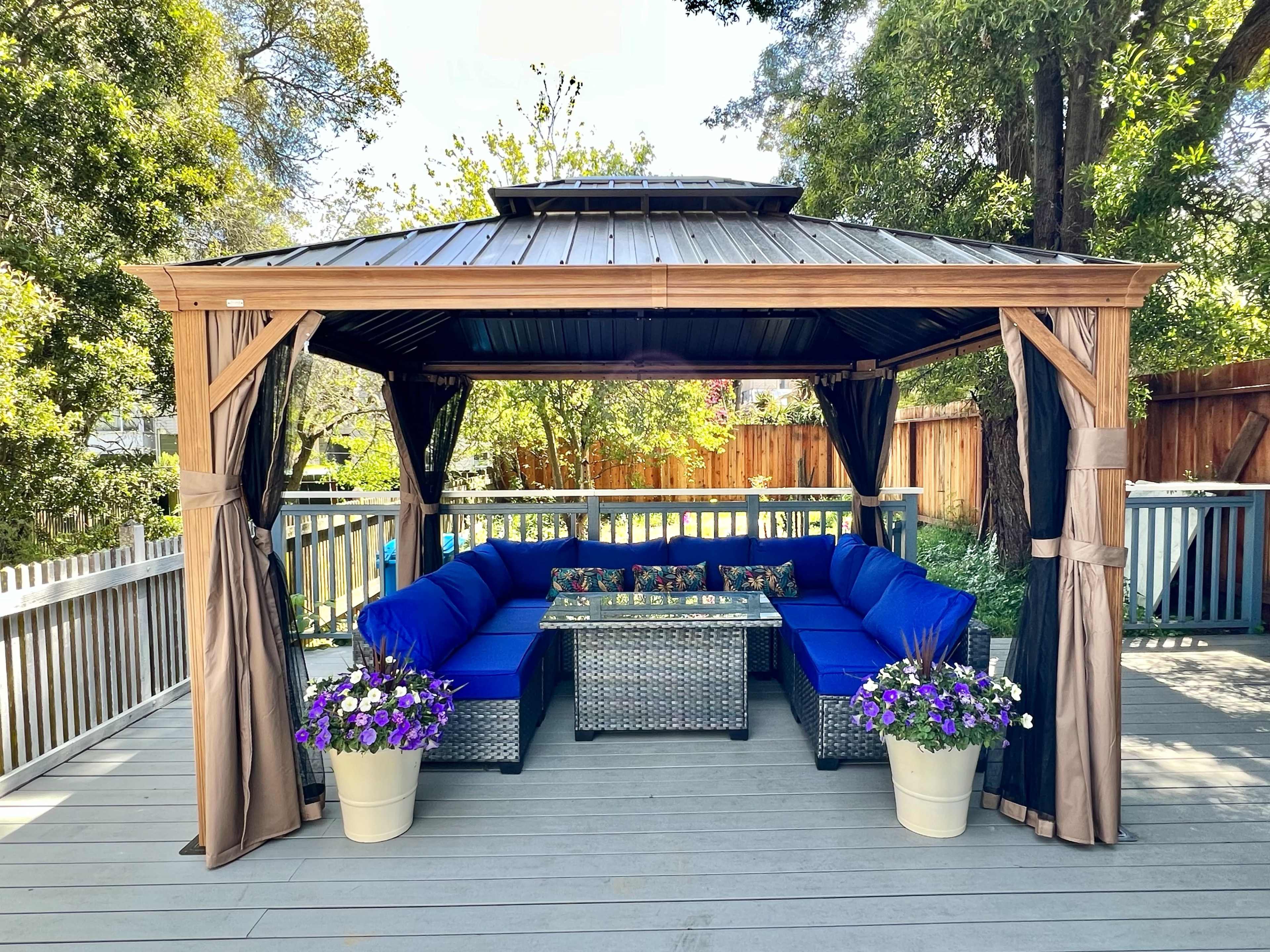A gazebo with blue upholstered seating surrounds a glass-top table, placed on a deck adorned with potted purple flowers.