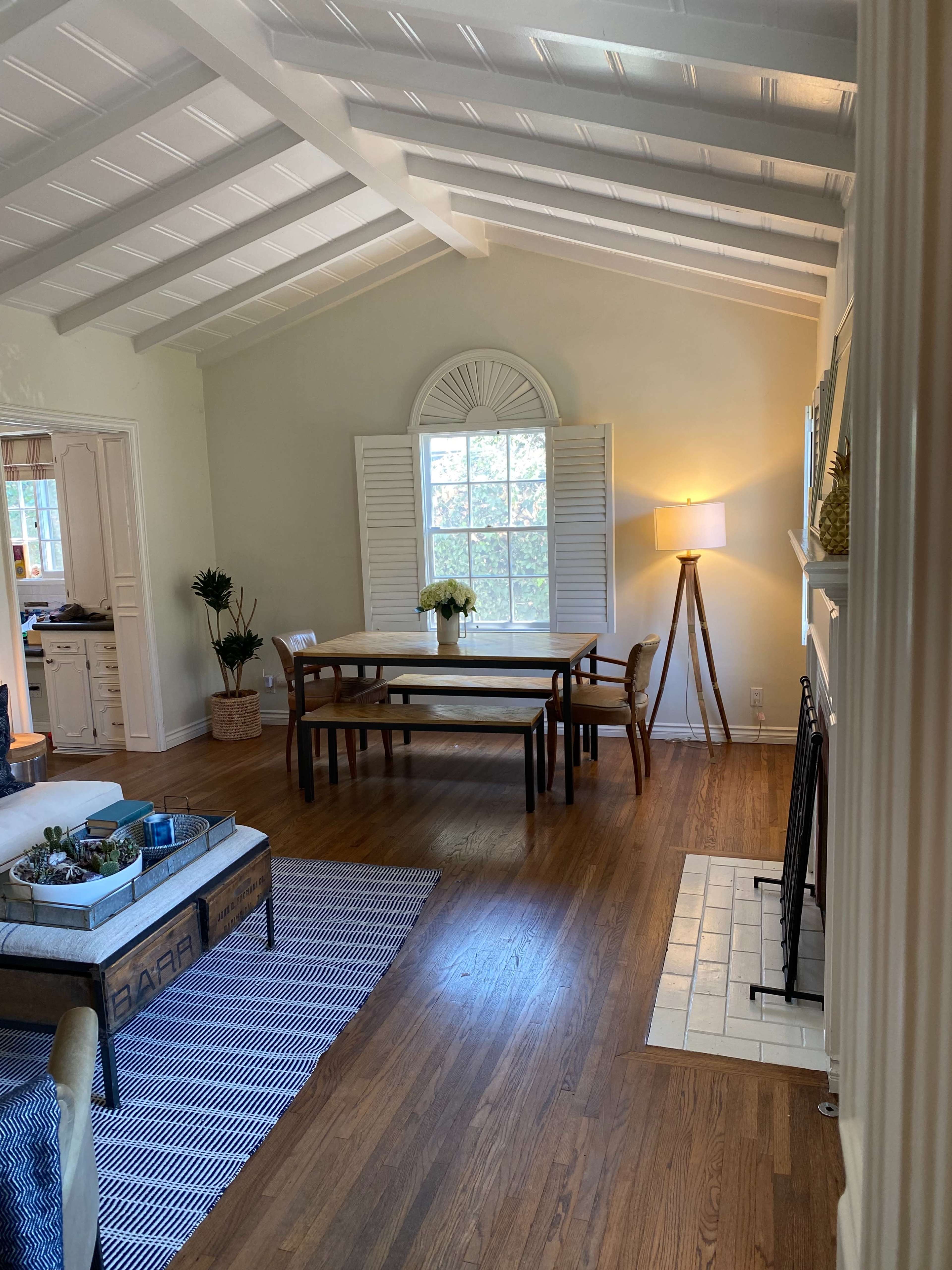 A spacious living room features a wooden dining table, a lamp, and a seating area with a coffee table, all illuminated by natural light from a window.