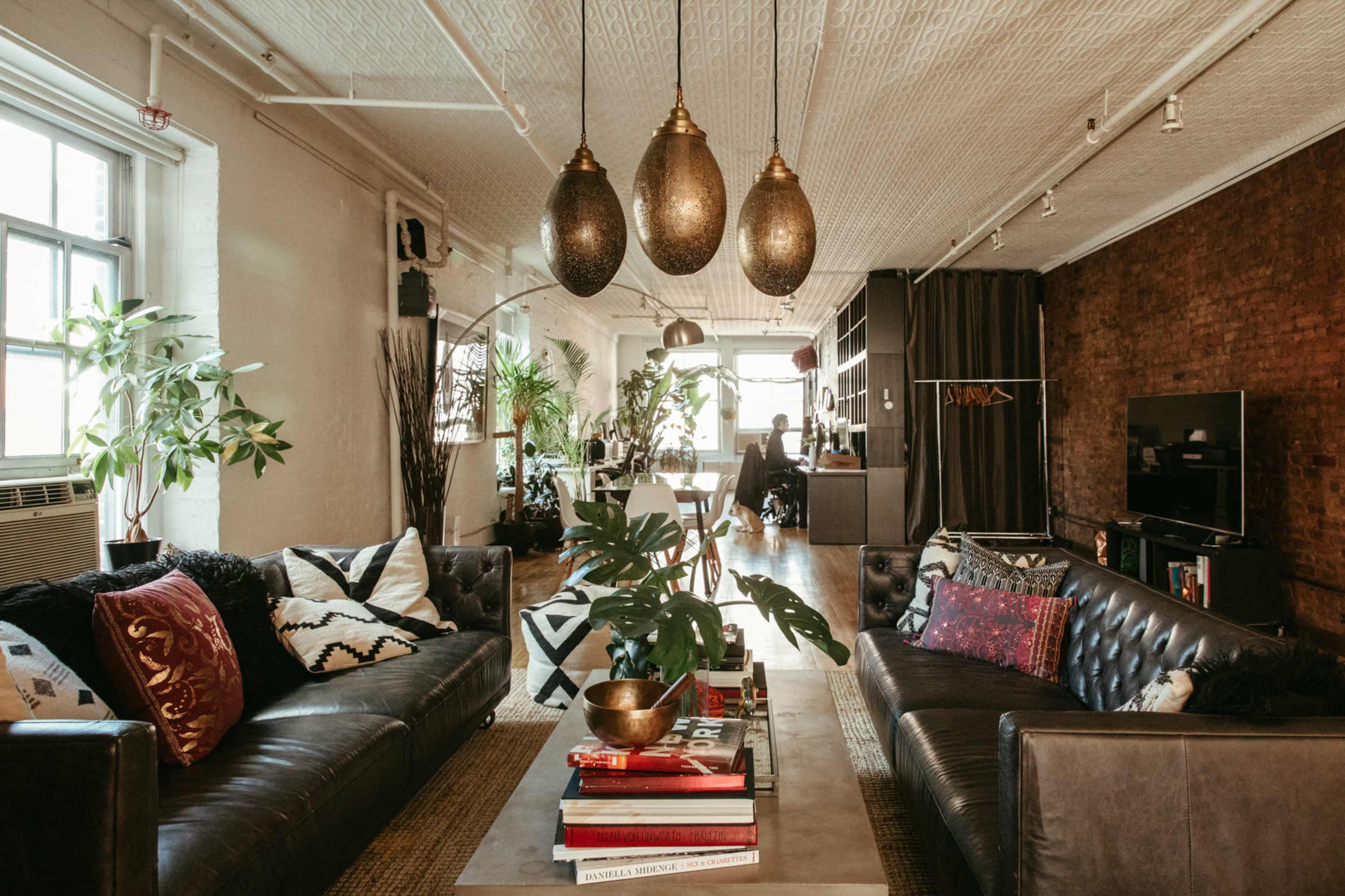 A stylish living room with a dark leather sofa, a central coffee table stacked with books, and large pendant lights hanging from the ceiling, alongside greenery and a modern TV setup against a brick wall.