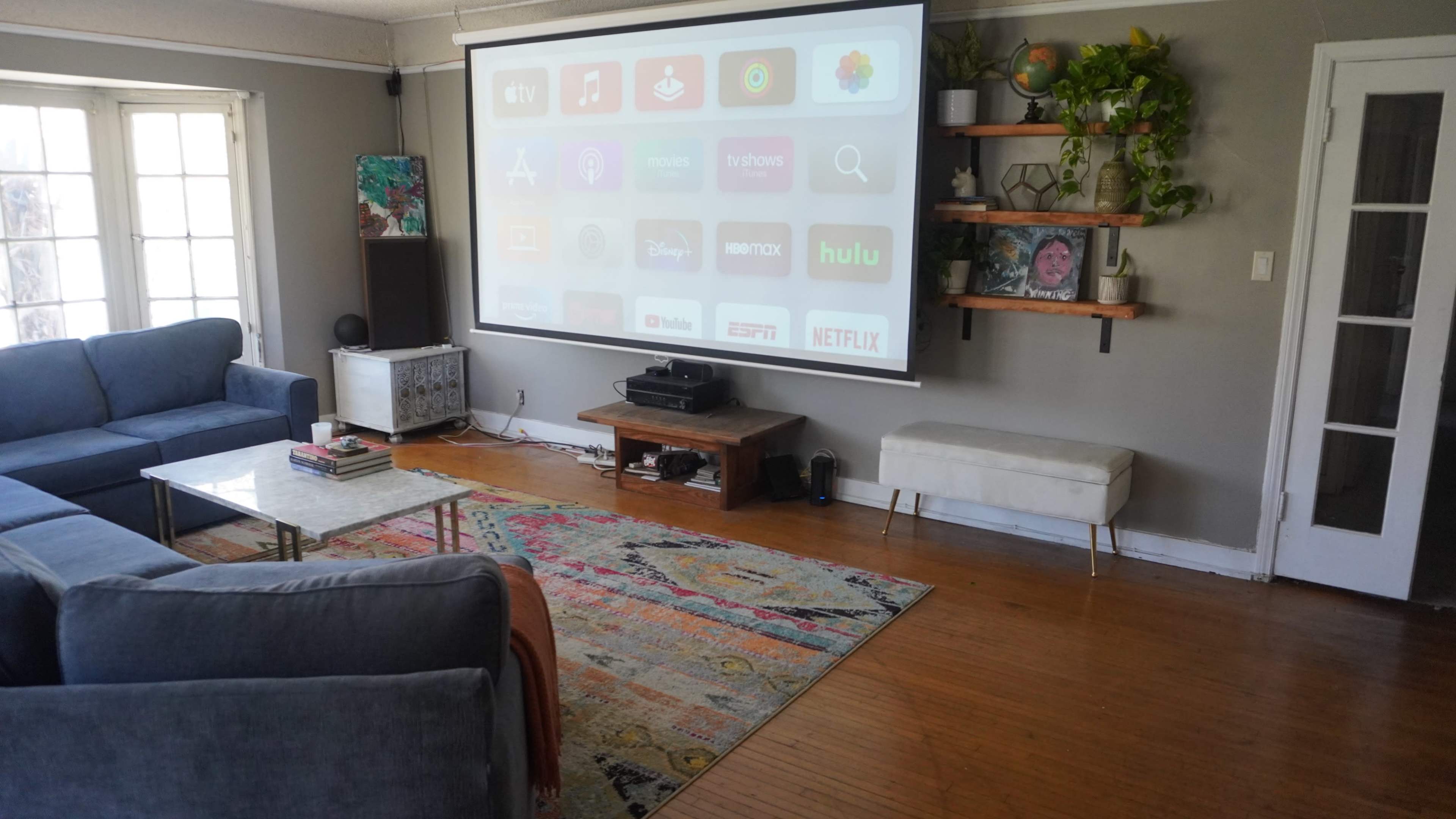 A living room features a large projection screen displaying various streaming services, with a sectional couch, a glass coffee table, and shelves adorned with plants and decor.