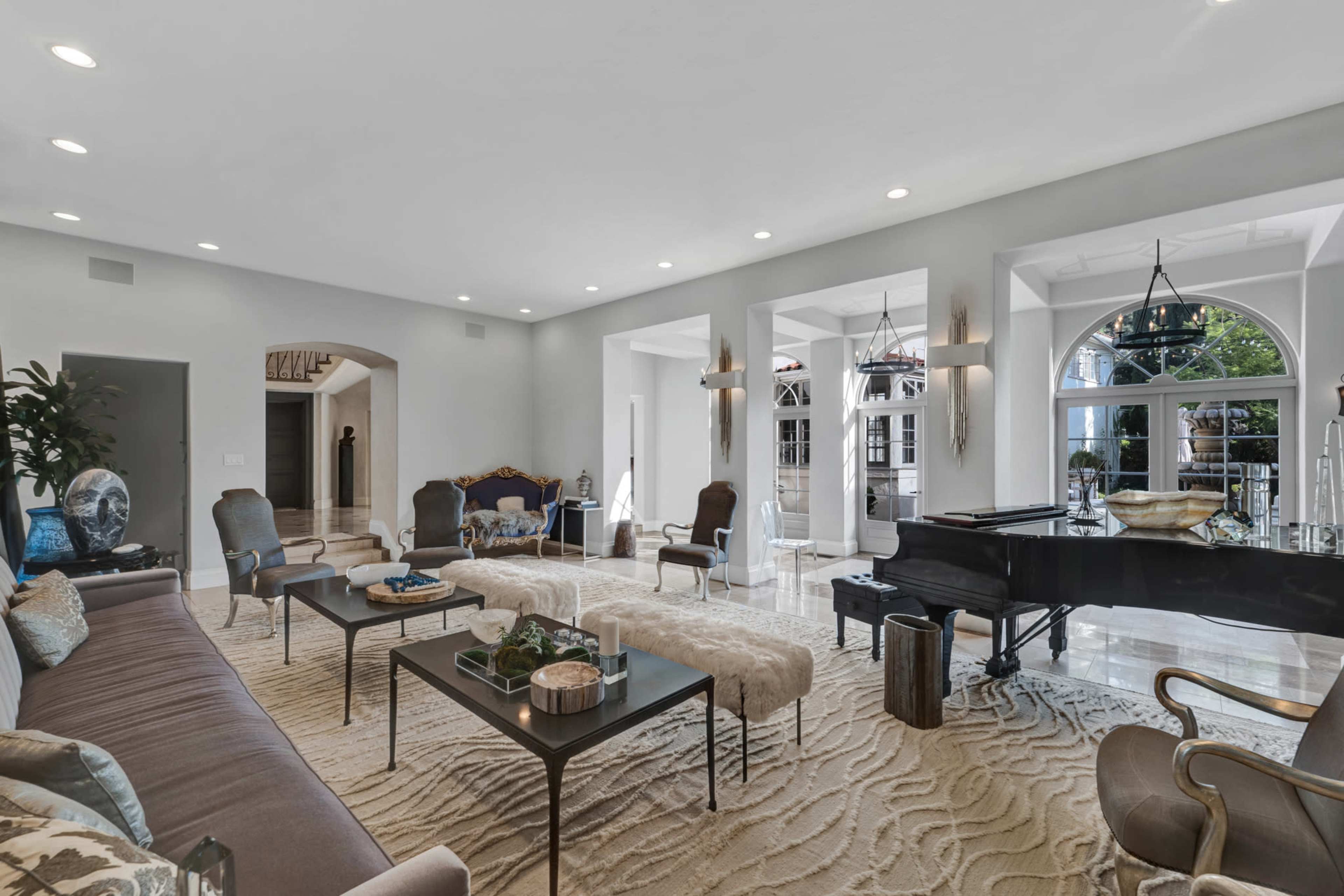 A spacious living room featuring a piano, an area rug, and a mix of seating arrangements, with large windows allowing natural light to enter.