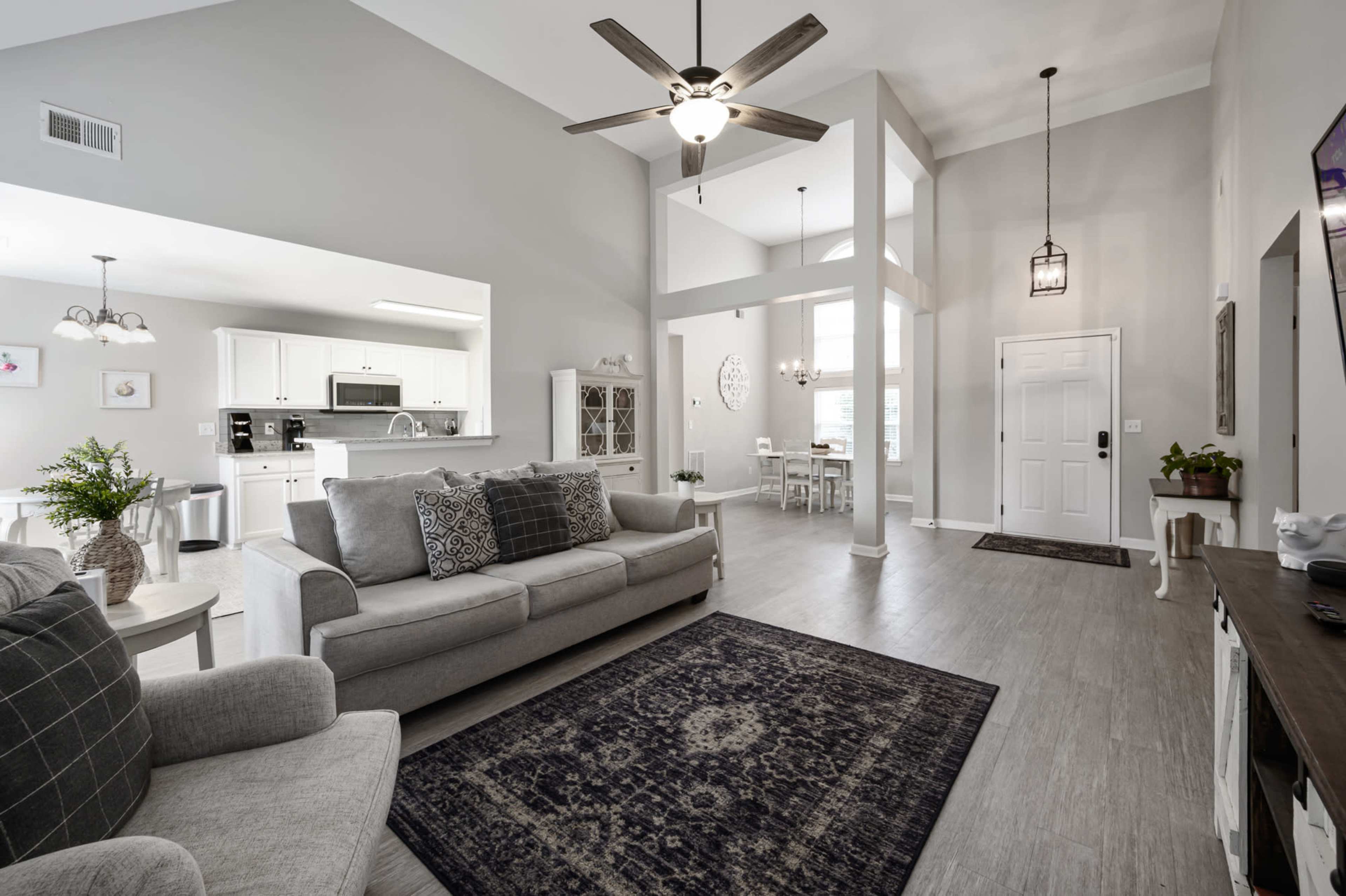 The image shows a bright, open living area featuring a light-colored sofa, a patterned area rug, and a dining space with a table in the background.