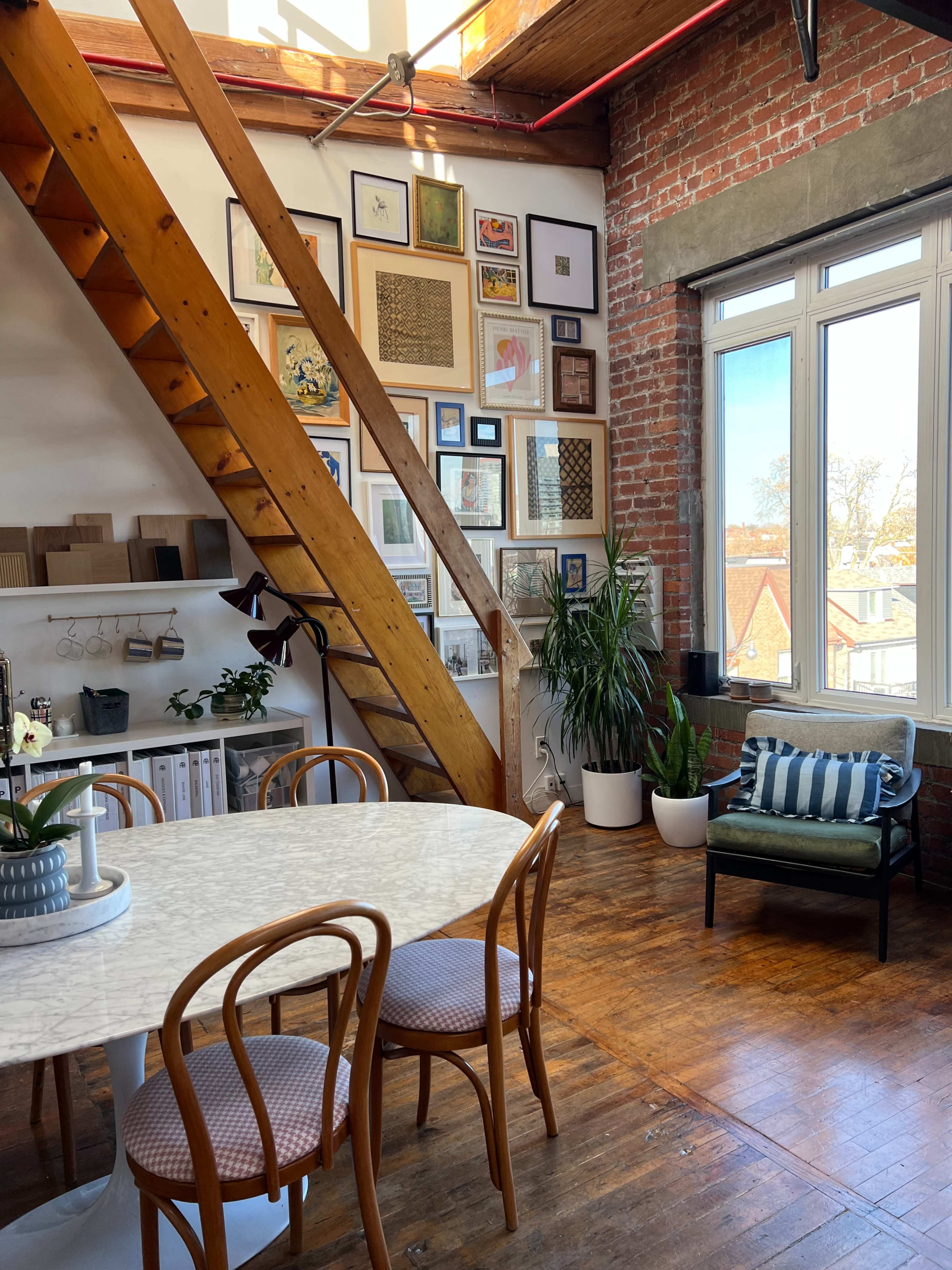 A bright room features a wooden staircase, a round marble table with four chairs, and a wall decorated with various framed artworks.