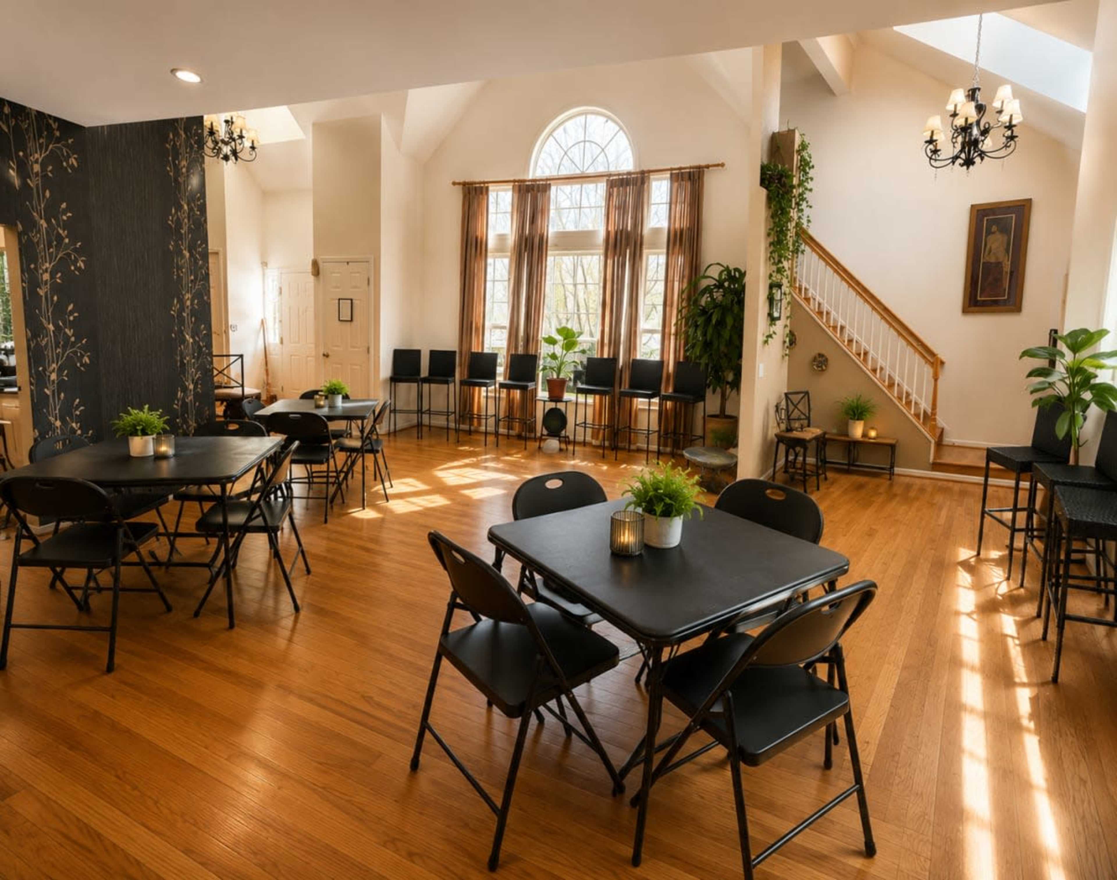 The image shows a spacious, well-lit room with wooden floors, multiple black folding tables and chairs, large windows, and decorative plants.