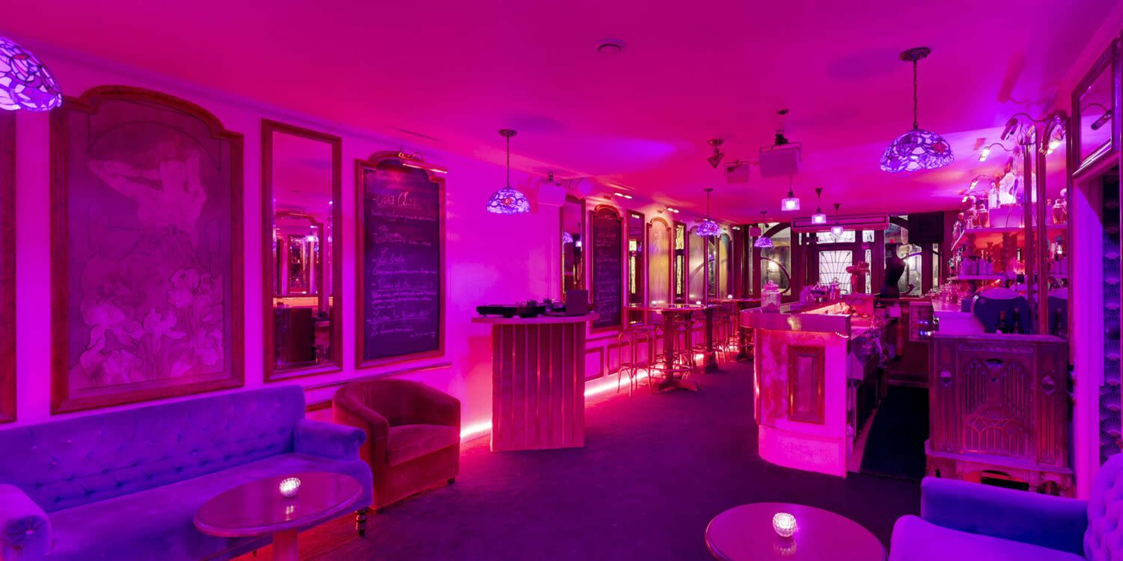 A dimly lit bar interior with purple lighting, featuring plush seating, a long bar with illuminated shelves, and decorative mirrors on the walls.