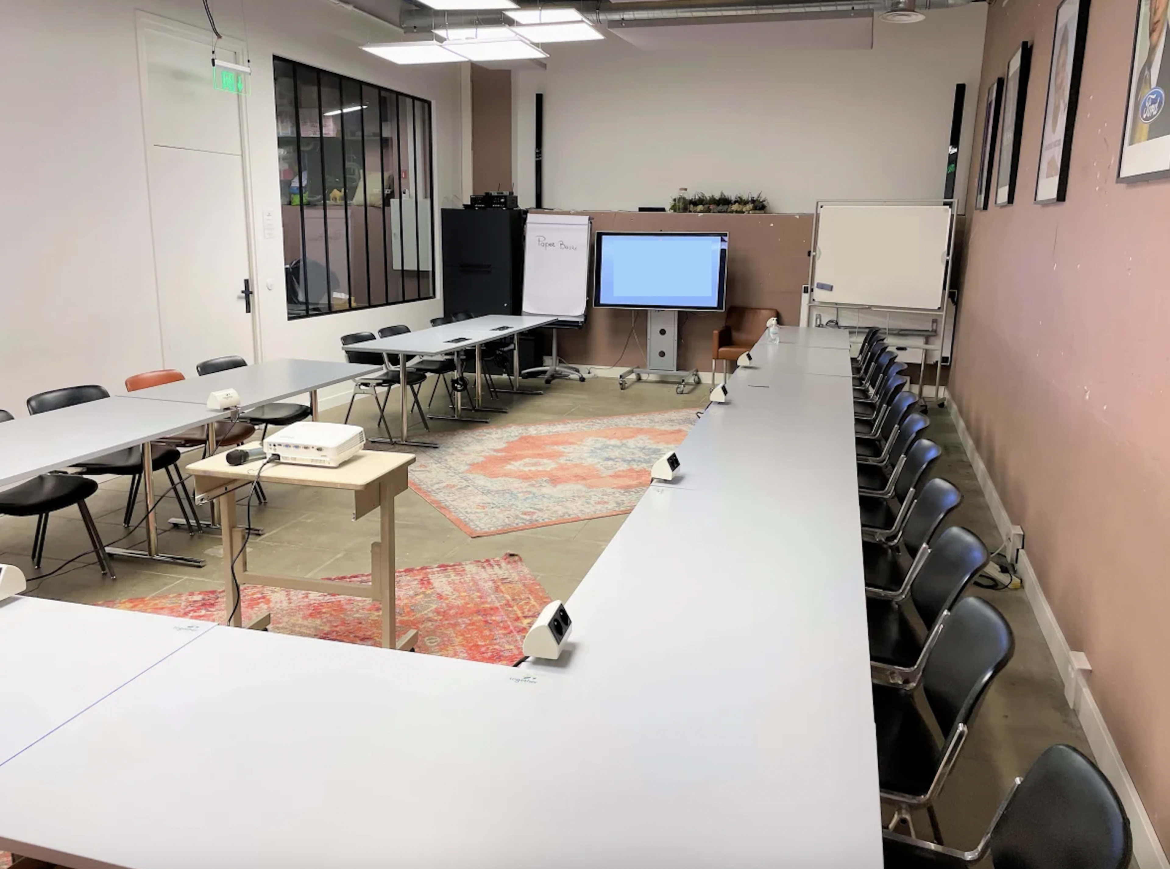 A conference room features two long tables arranged in a U-shape, with black chairs aligned along the sides, a projector, and a large screen at one end.