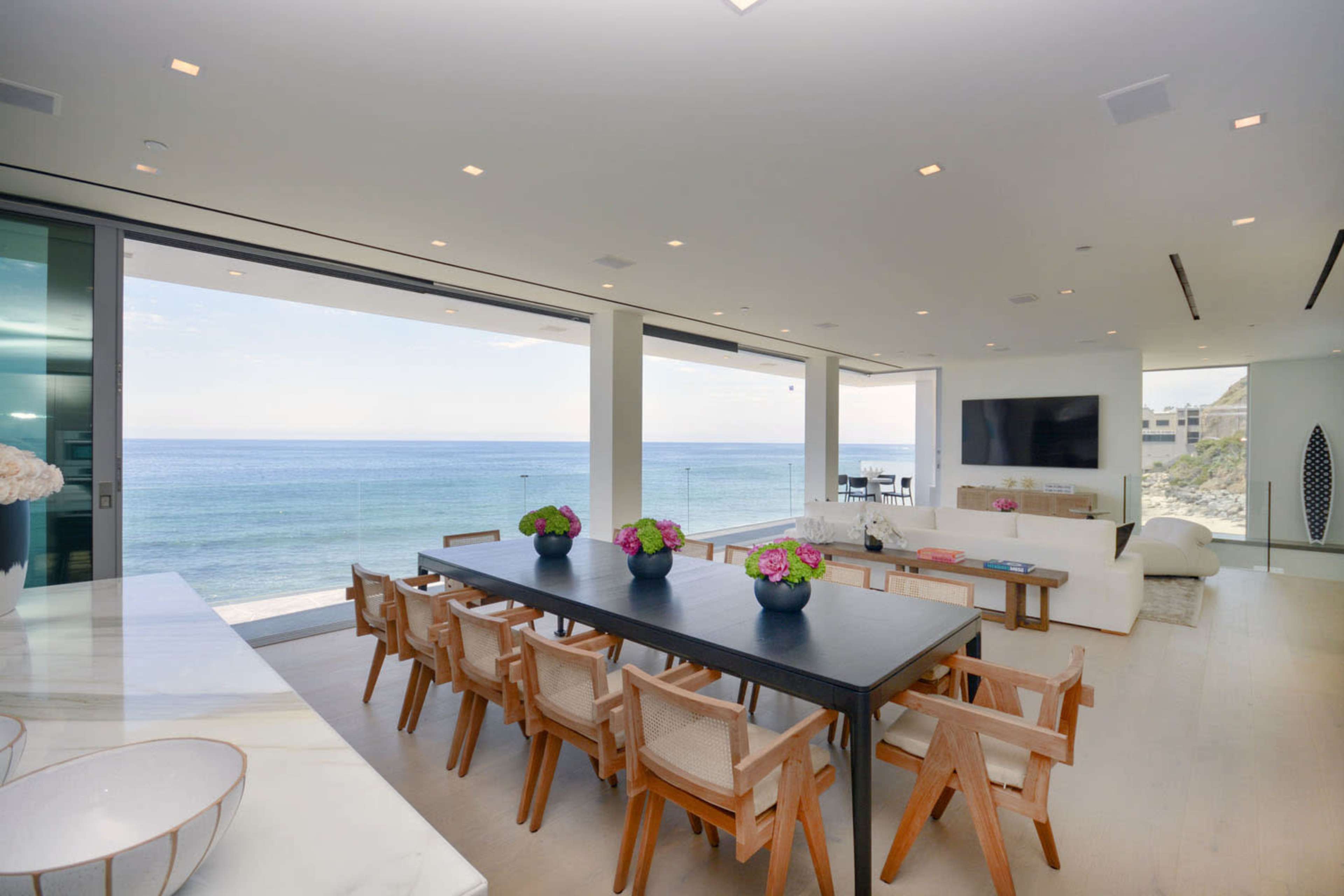 A modern dining area features a long black table surrounded by wooden chairs, with large windows offering a view of the ocean.