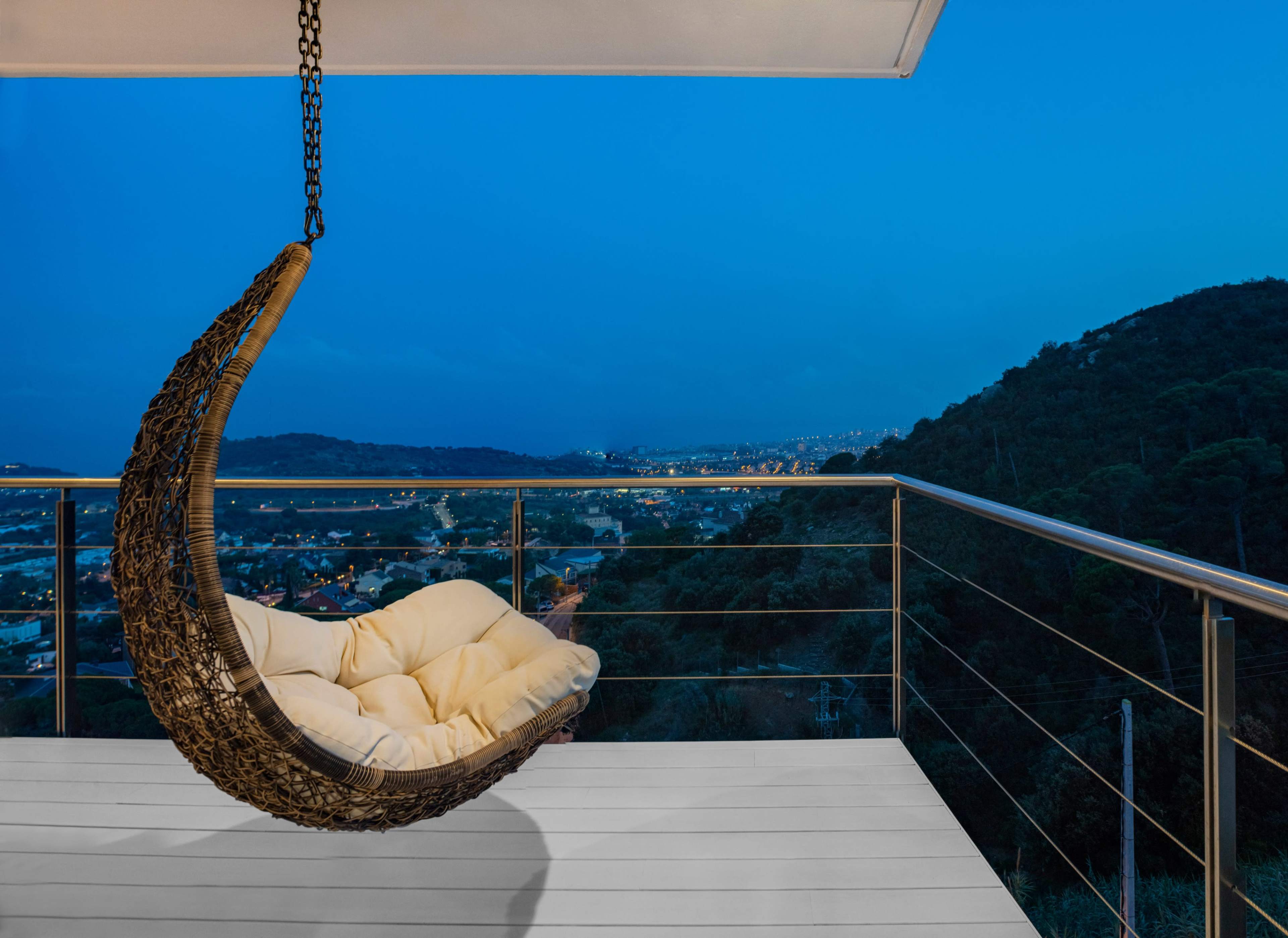 A hanging chair with a cushion is positioned on a balcony overlooking a cityscape and hills at dusk.