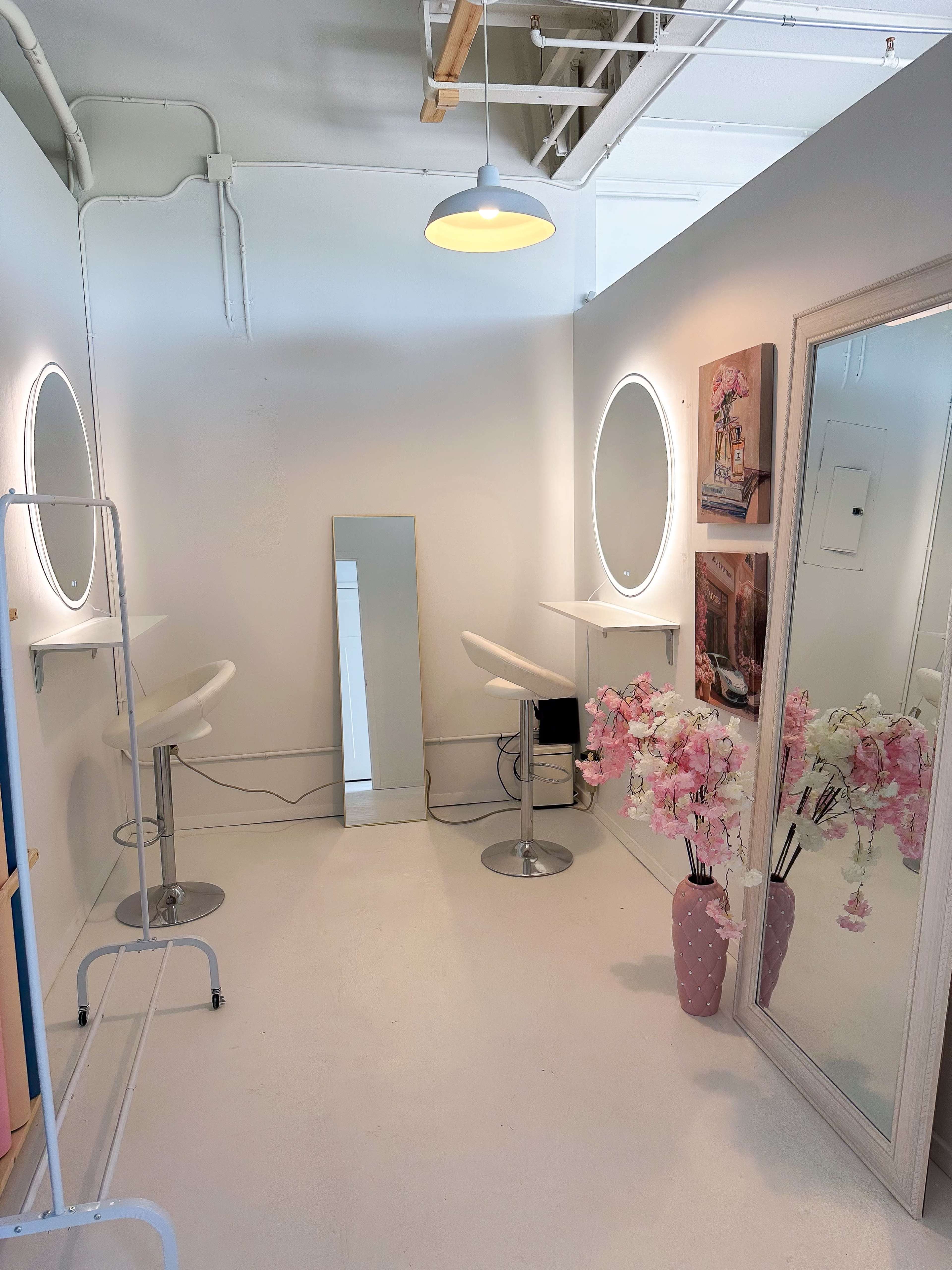 The image shows a light-colored salon space with two white bar stools, a full-length mirror, a flower arrangement in a pink vase, and framed artwork on the walls.