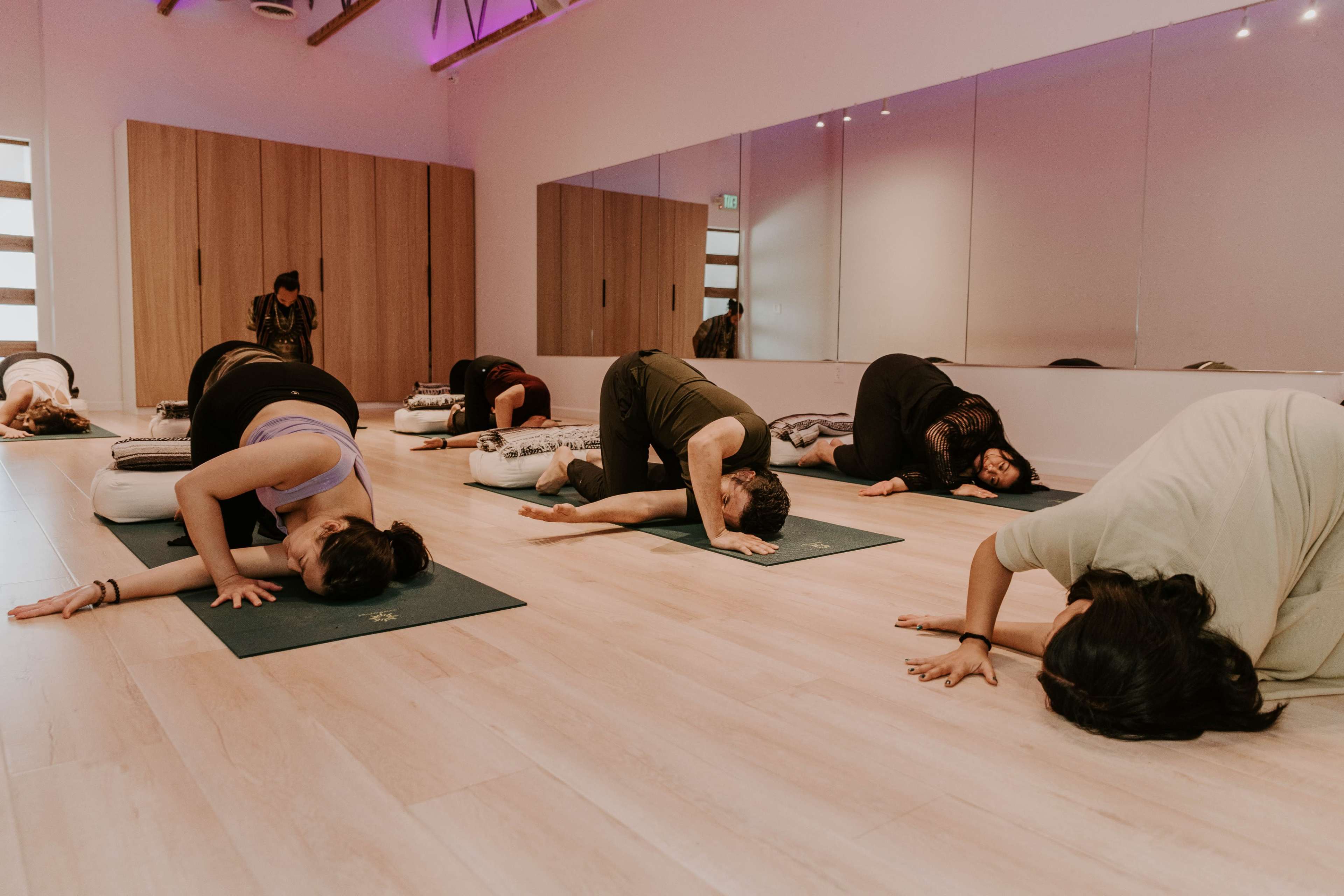 A group of people are practicing yoga on mats in a brightly lit room with wooden floors and mirrors.