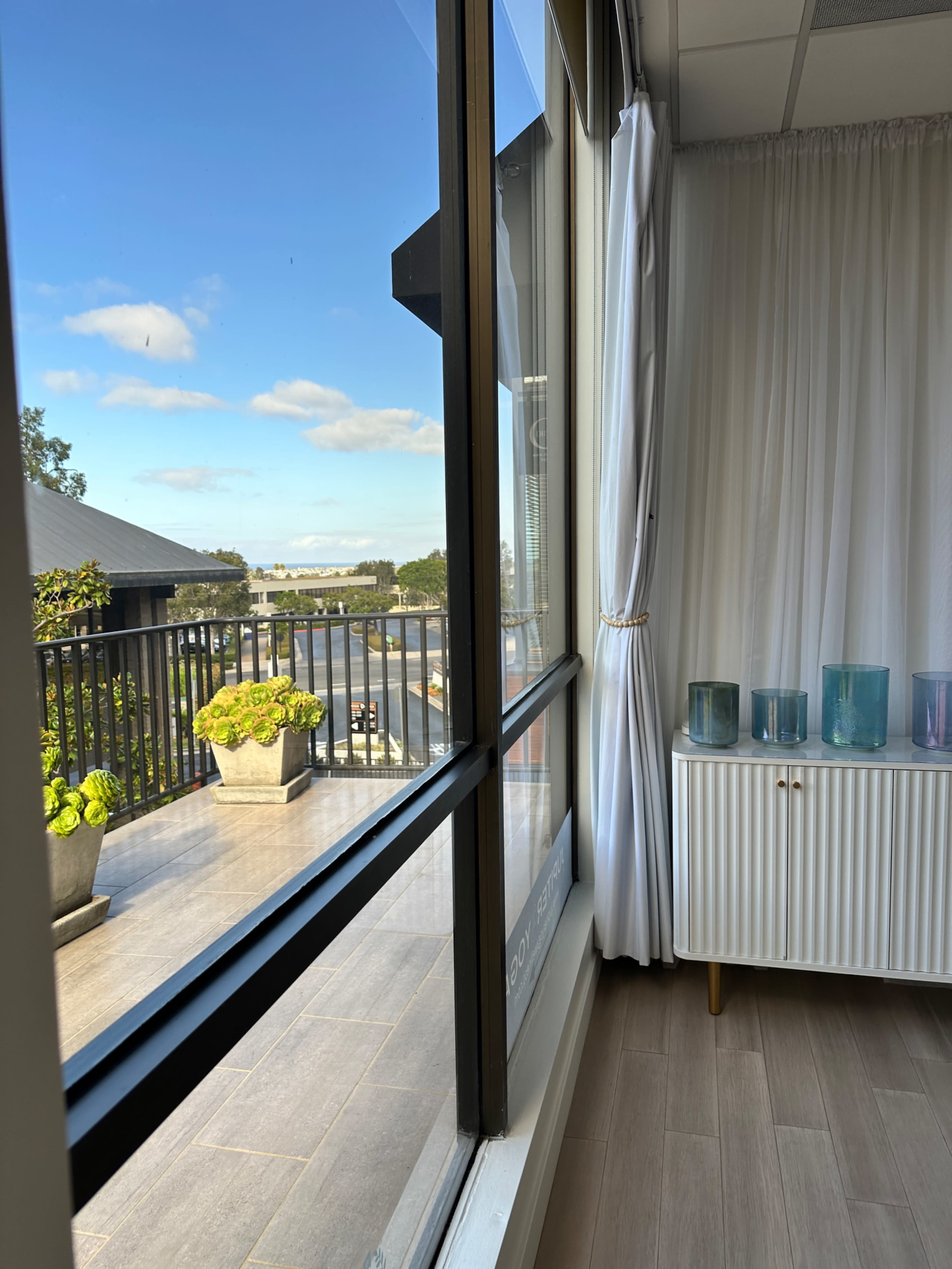 The image shows a bright interior space with a window revealing a balcony area featuring potted plants and a scenic view outside.
