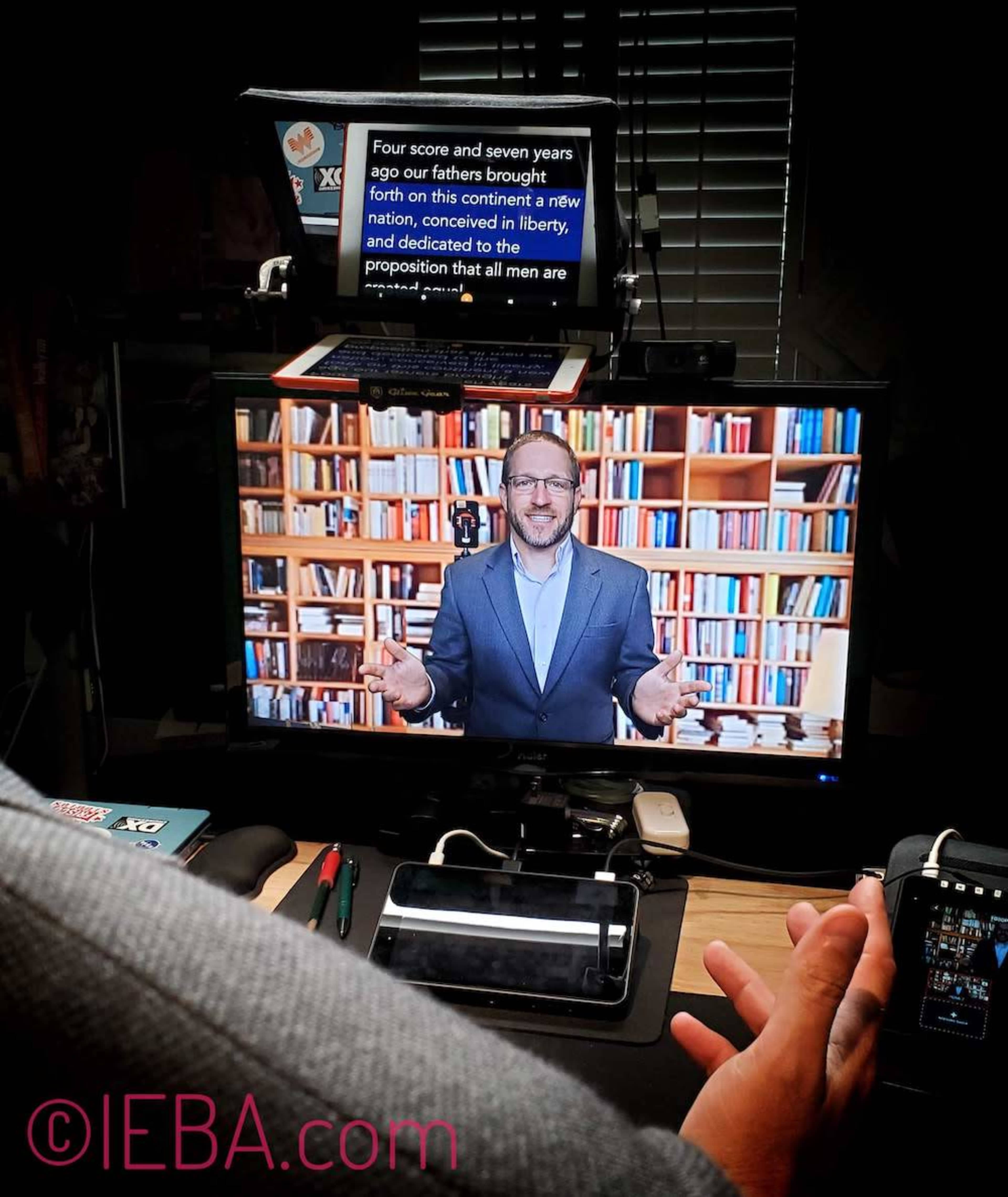 A person is presenting from a computer screen in front of a backdrop of bookshelves, while a caption displays text above the monitor.