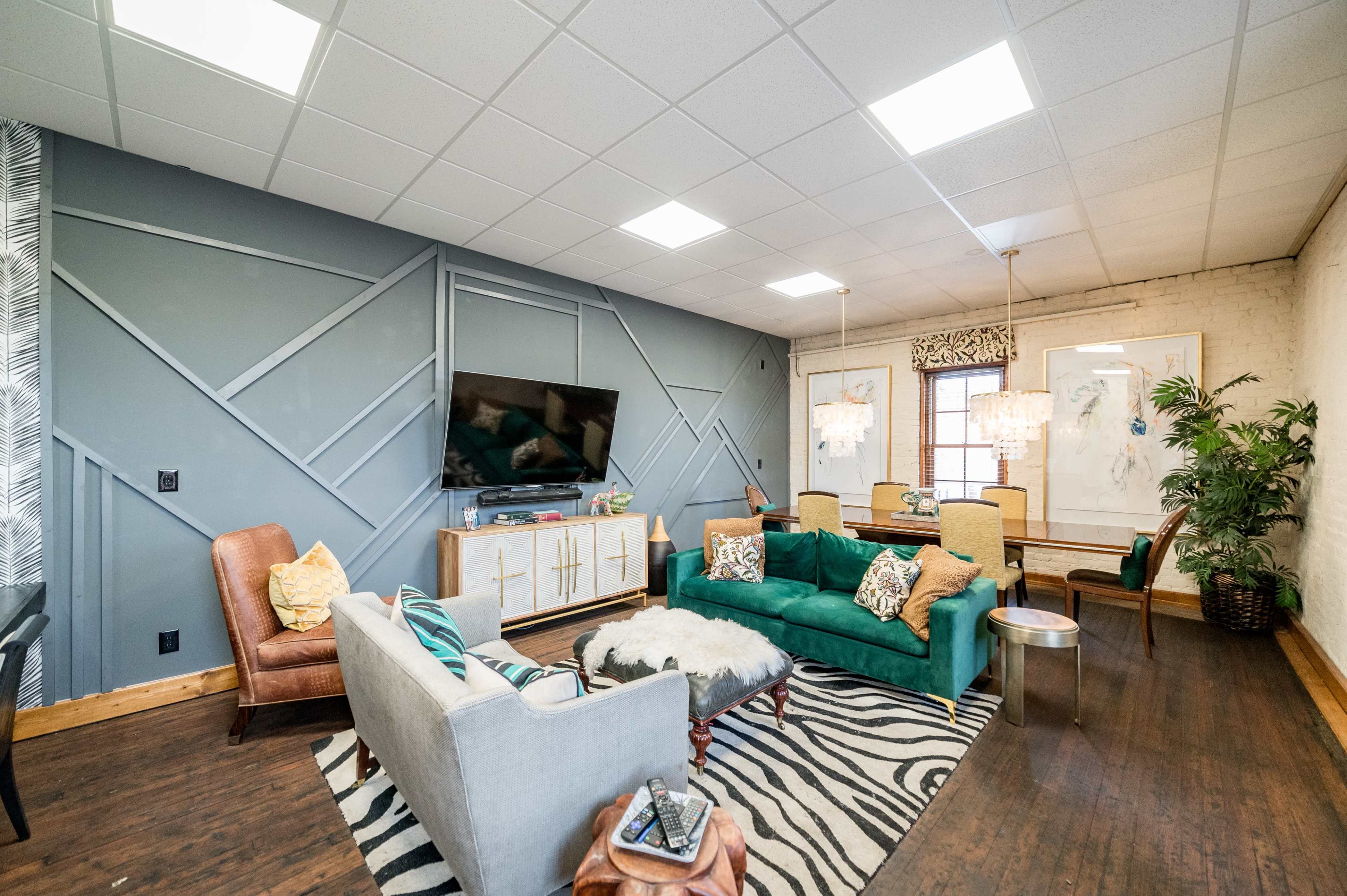 A modern living space featuring a mix of seating options, including a light gray sofa, a tan armchair, and a patterned rug, with a large television mounted on the wall and decorative lighting fixtures.