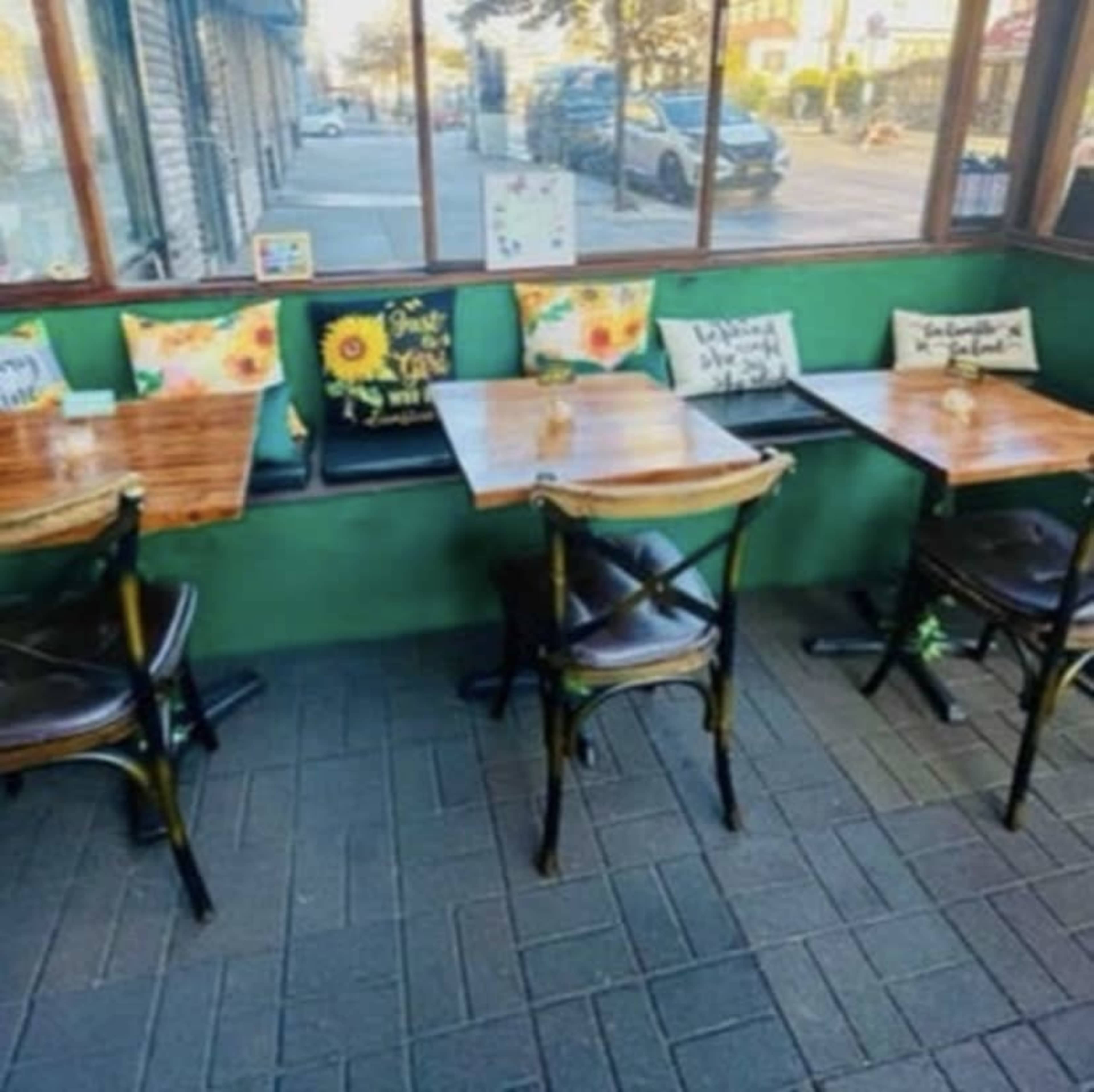 A café interior with wooden tables and chairs arranged beside a window, featuring colorful cushions along the wall.