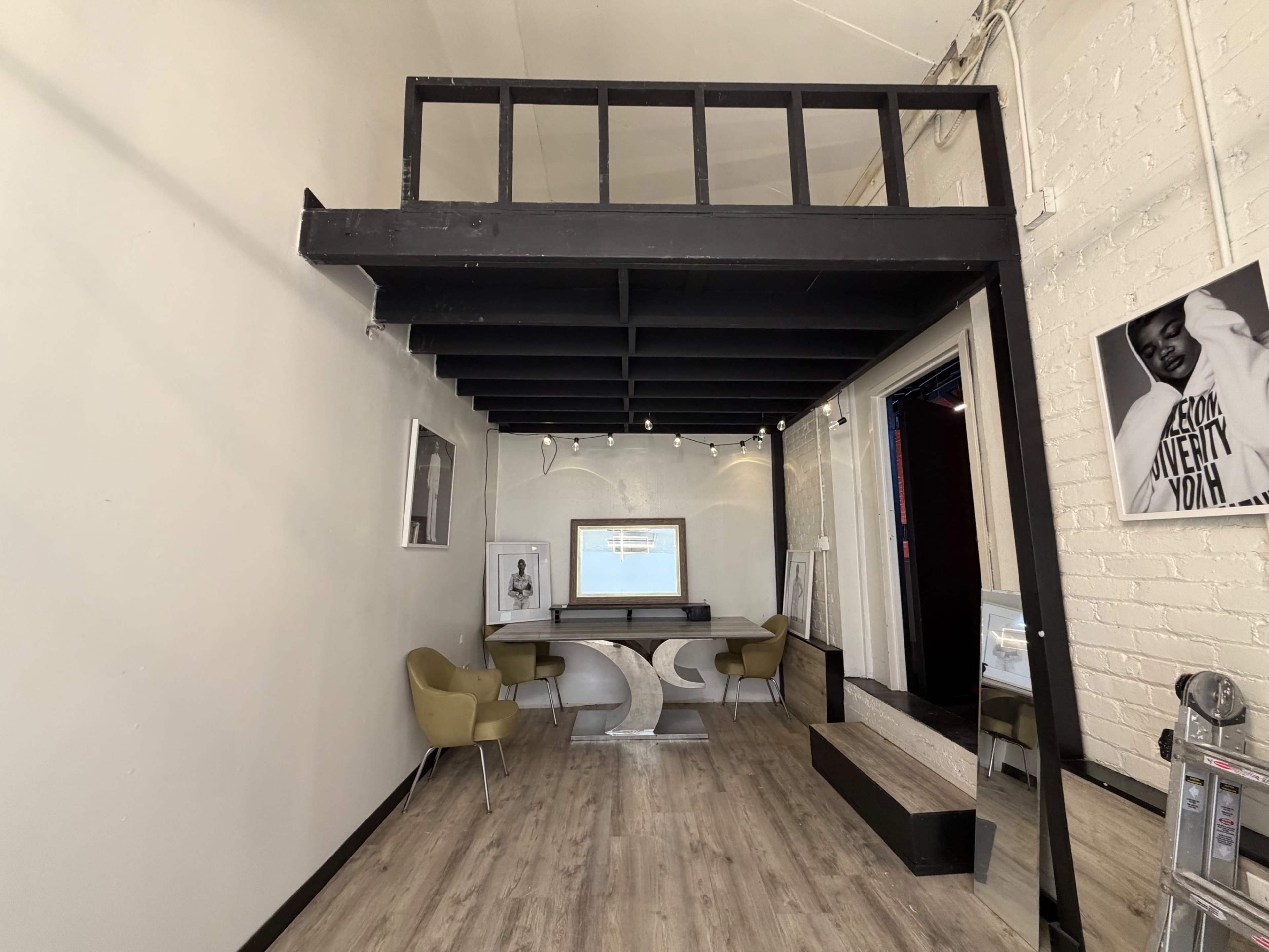 The image shows a minimalist interior space with a small loft area, a curved table in the center, yellow chairs, and a large mirror along one wall.