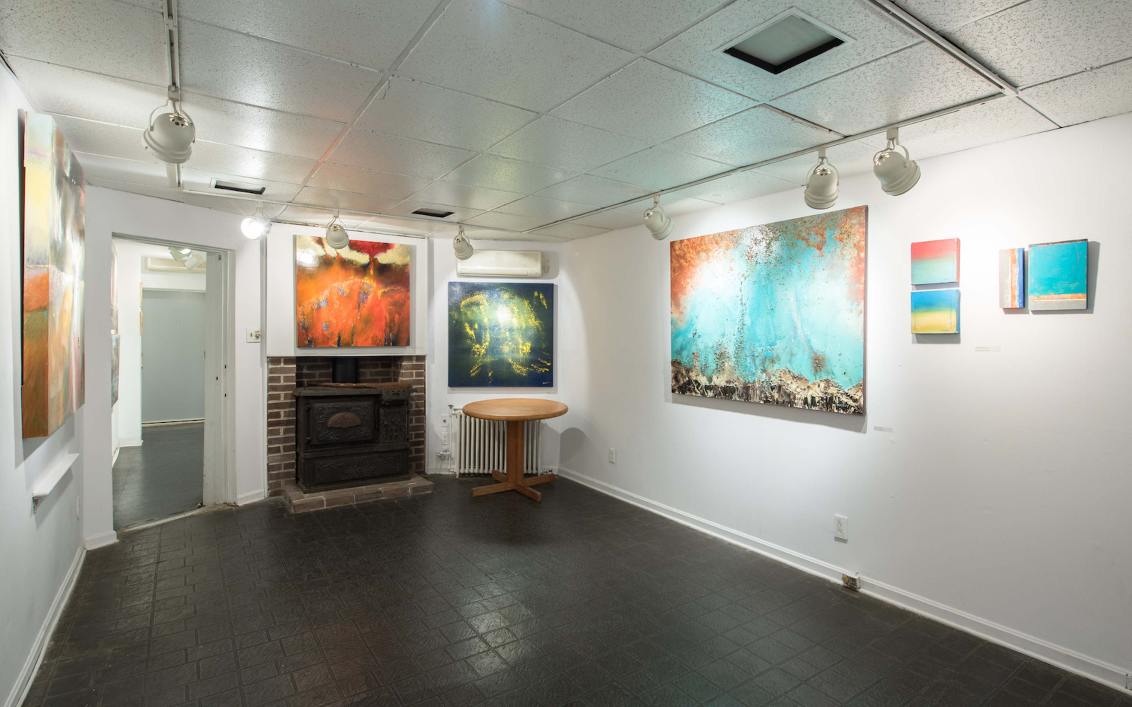 An art gallery with several colorful paintings displayed on white walls, featuring a round wooden table in the center and a brick fireplace along one wall.