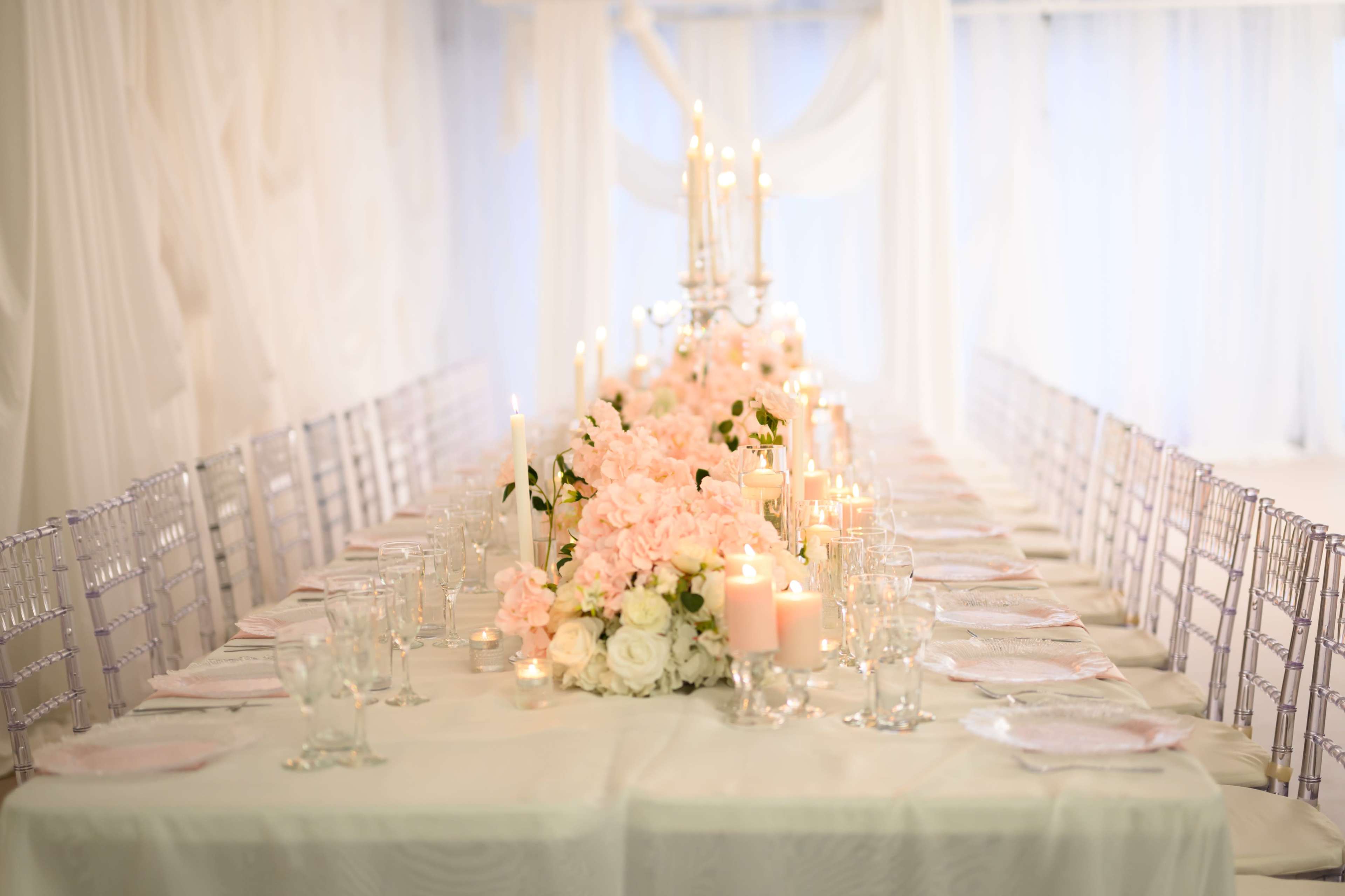 A long banquet table is elegantly arranged with white tablecloths, crystal chairs, floral centerpieces, candles, and place settings.