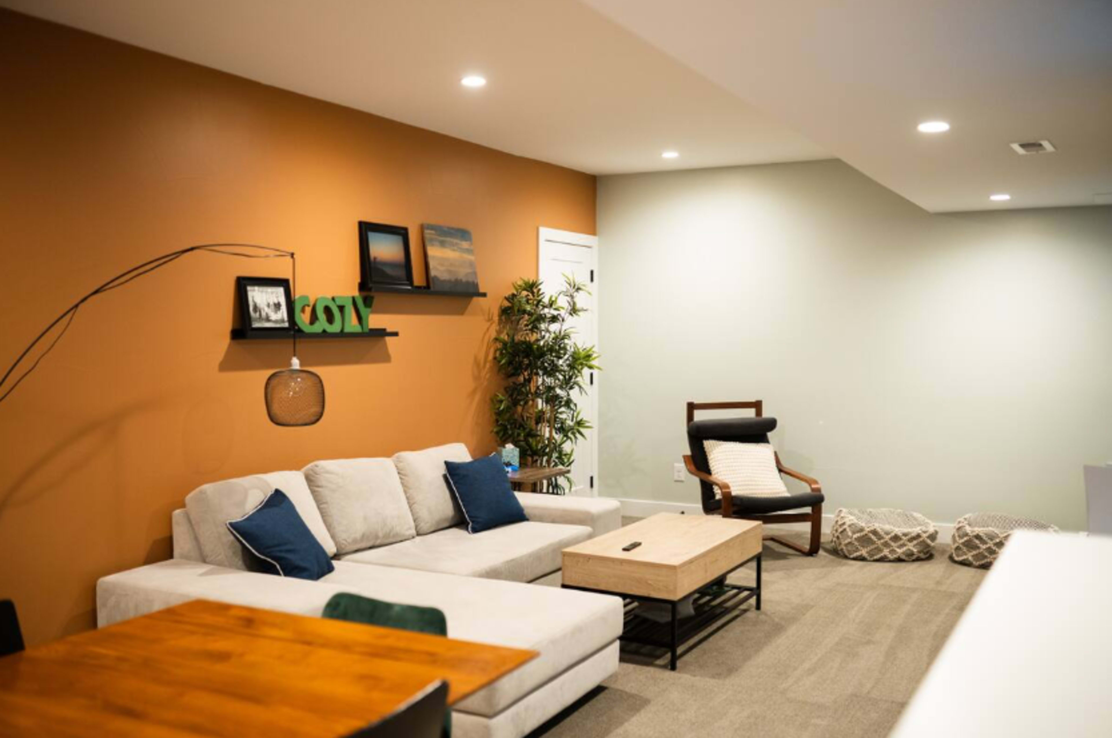 A modern living room features a light-colored sectional sofa, a wooden coffee table, framed pictures on the wall, and a potted plant in one corner.