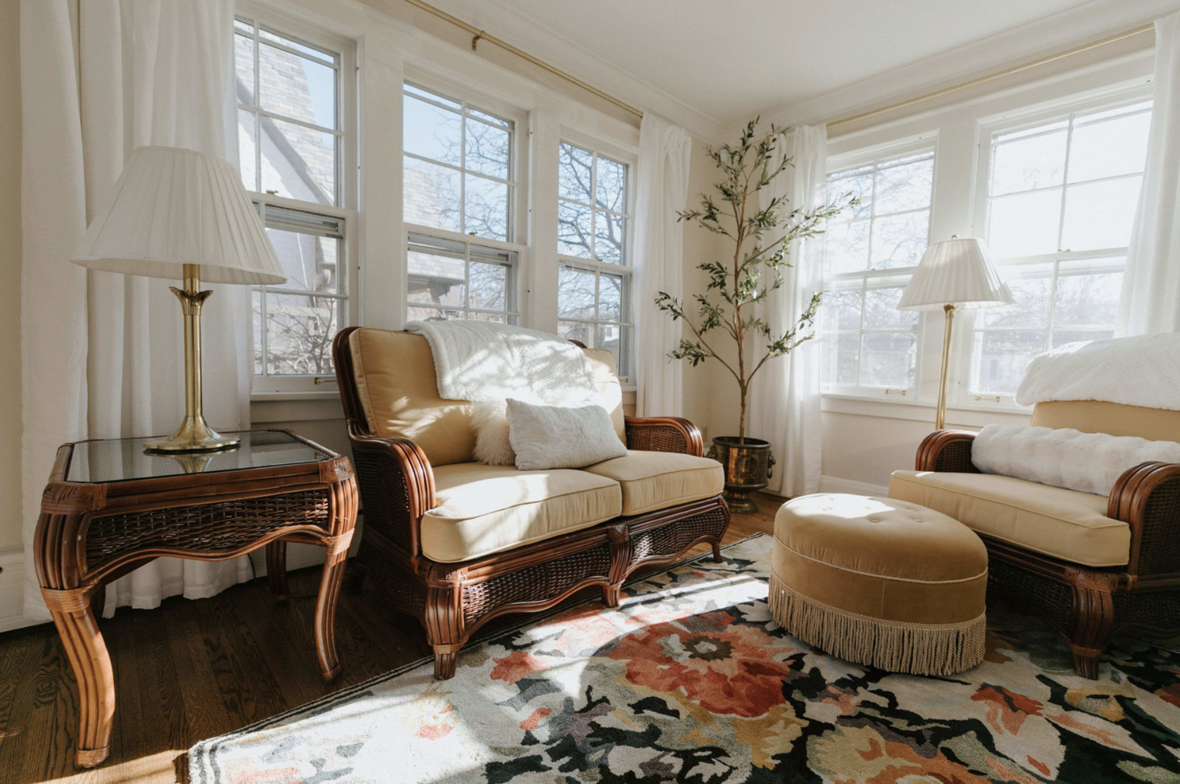 A bright living room features two tan upholstered chairs, a round ottoman with fringe, and a side table, all illuminated by natural light from large windows.