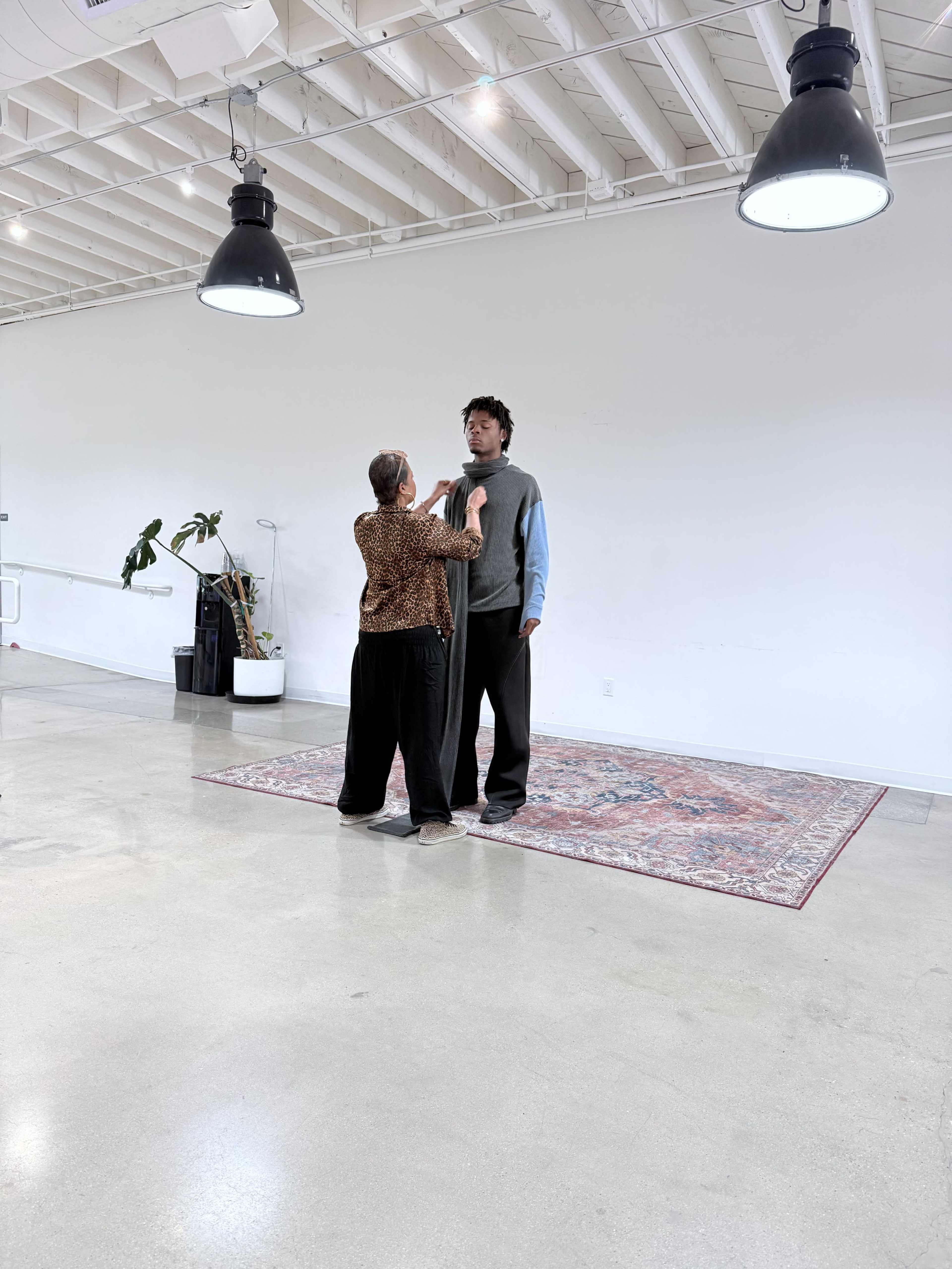 A woman in a patterned top and wide-legged pants interacts with a tall man wearing layered clothing in a spacious, well-lit room with a large rug on the floor.