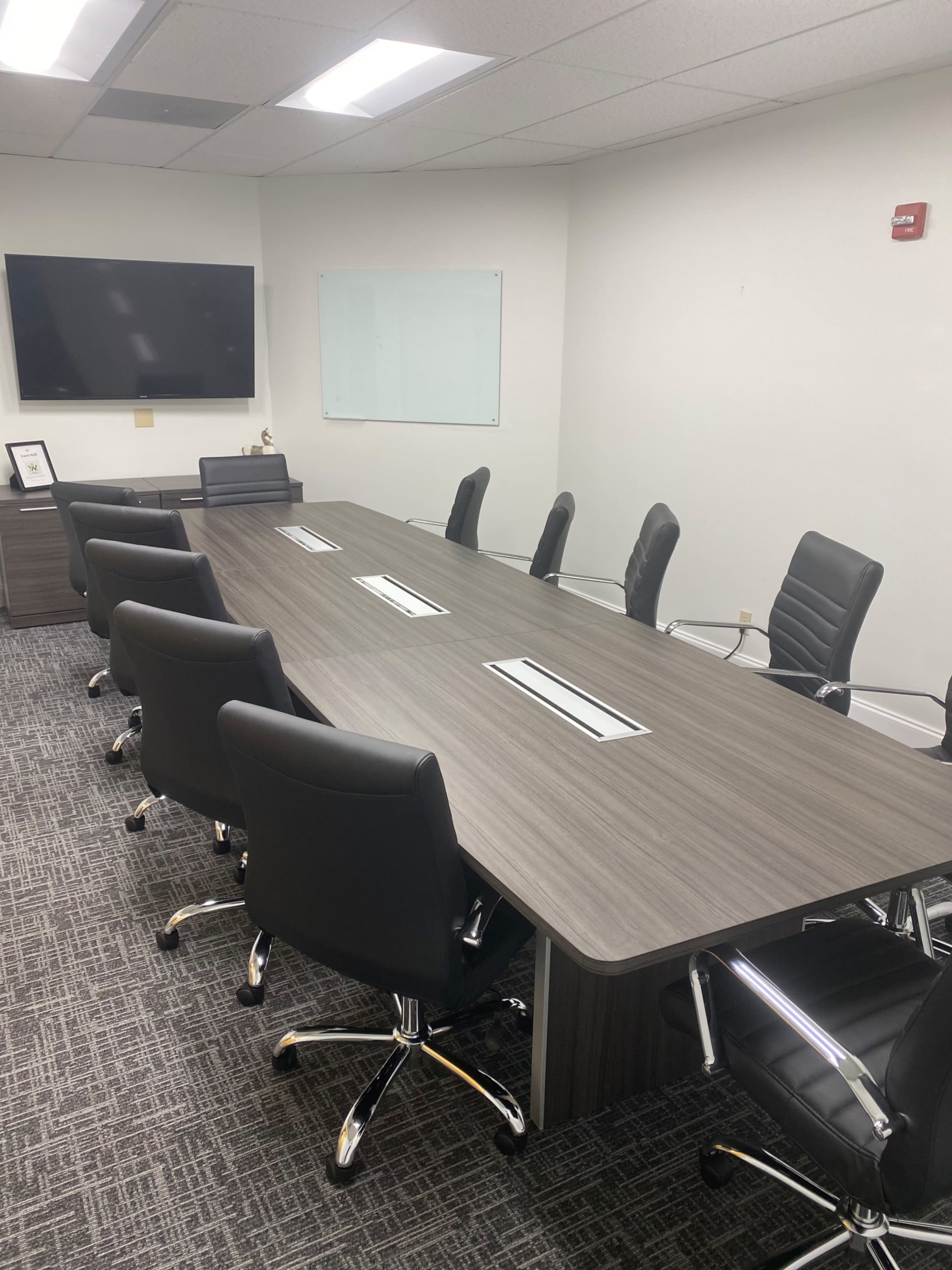 A large conference room features a long rectangular table surrounded by black ergonomic chairs, with a wall-mounted screen and a whiteboard.