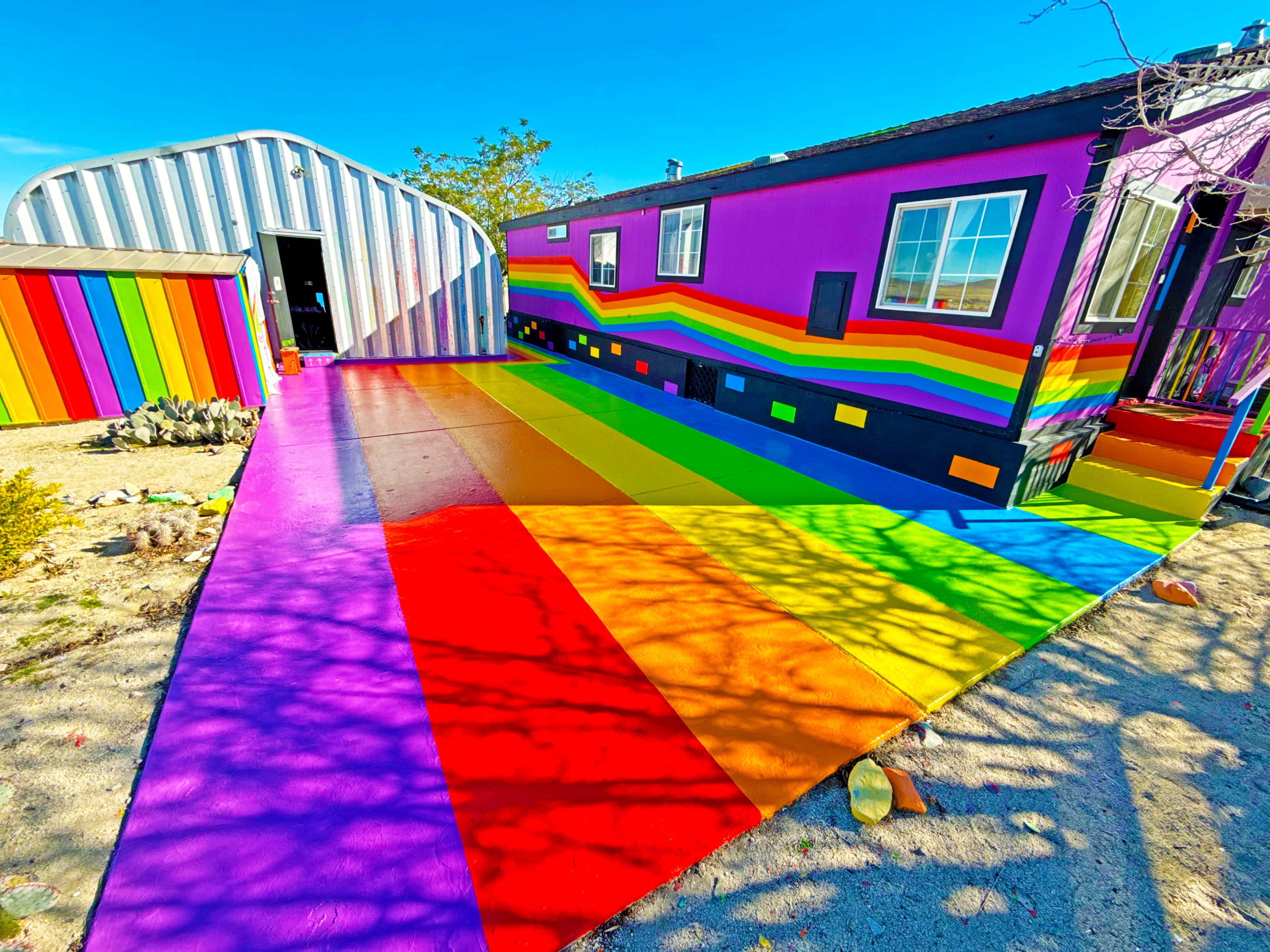 The image shows a colorful, rainbow-striped pathway leading to a brightly painted mobile home and a nearby shed.