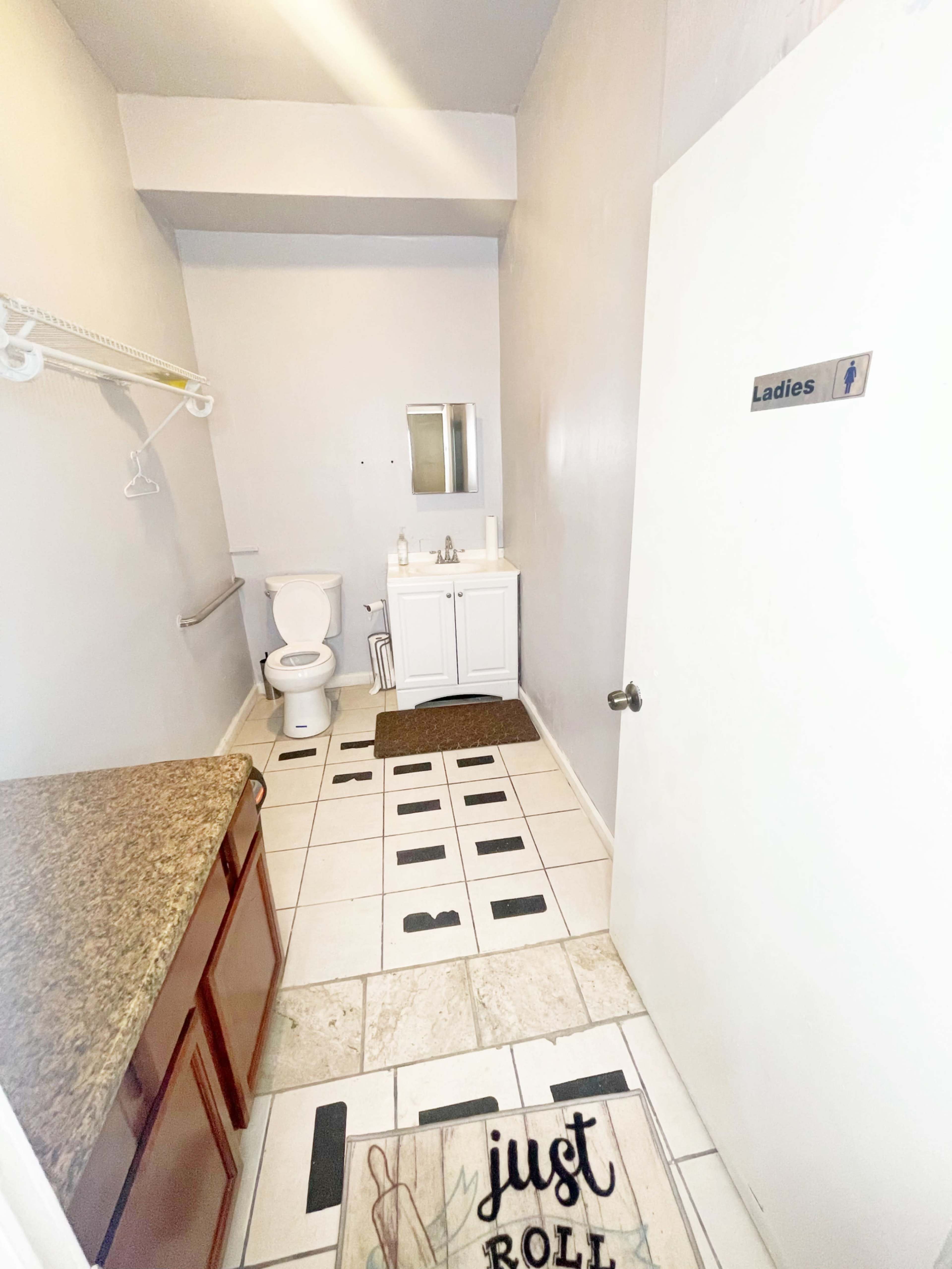A bathroom with a toilet, a sink, a mirror, and a cabinet, featuring tiled floors and a sign indicating it is a ladies' restroom.