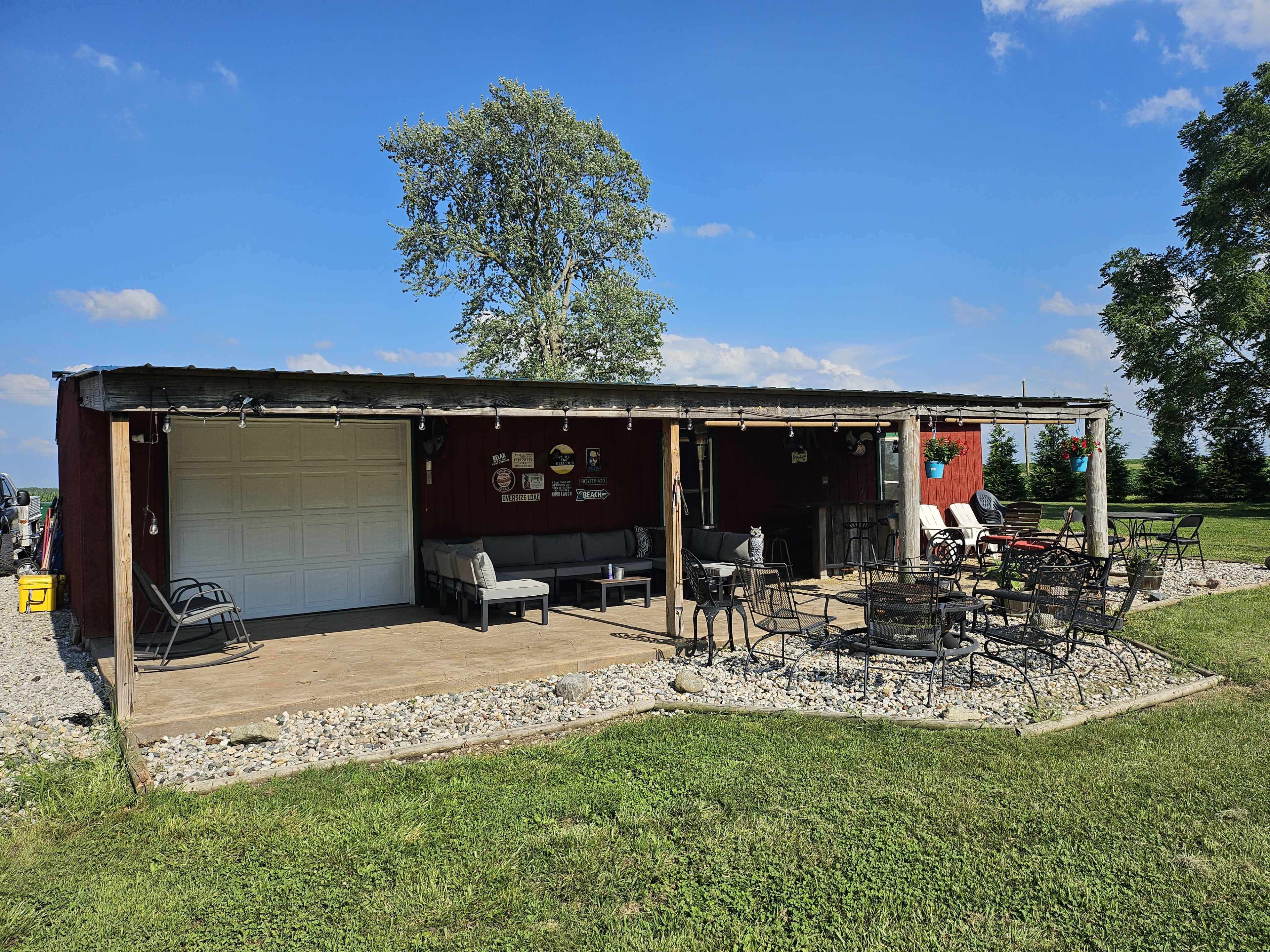 A rustic outdoor patio area with a covered seating space, surrounded by a grassy lawn and trees under a blue sky.