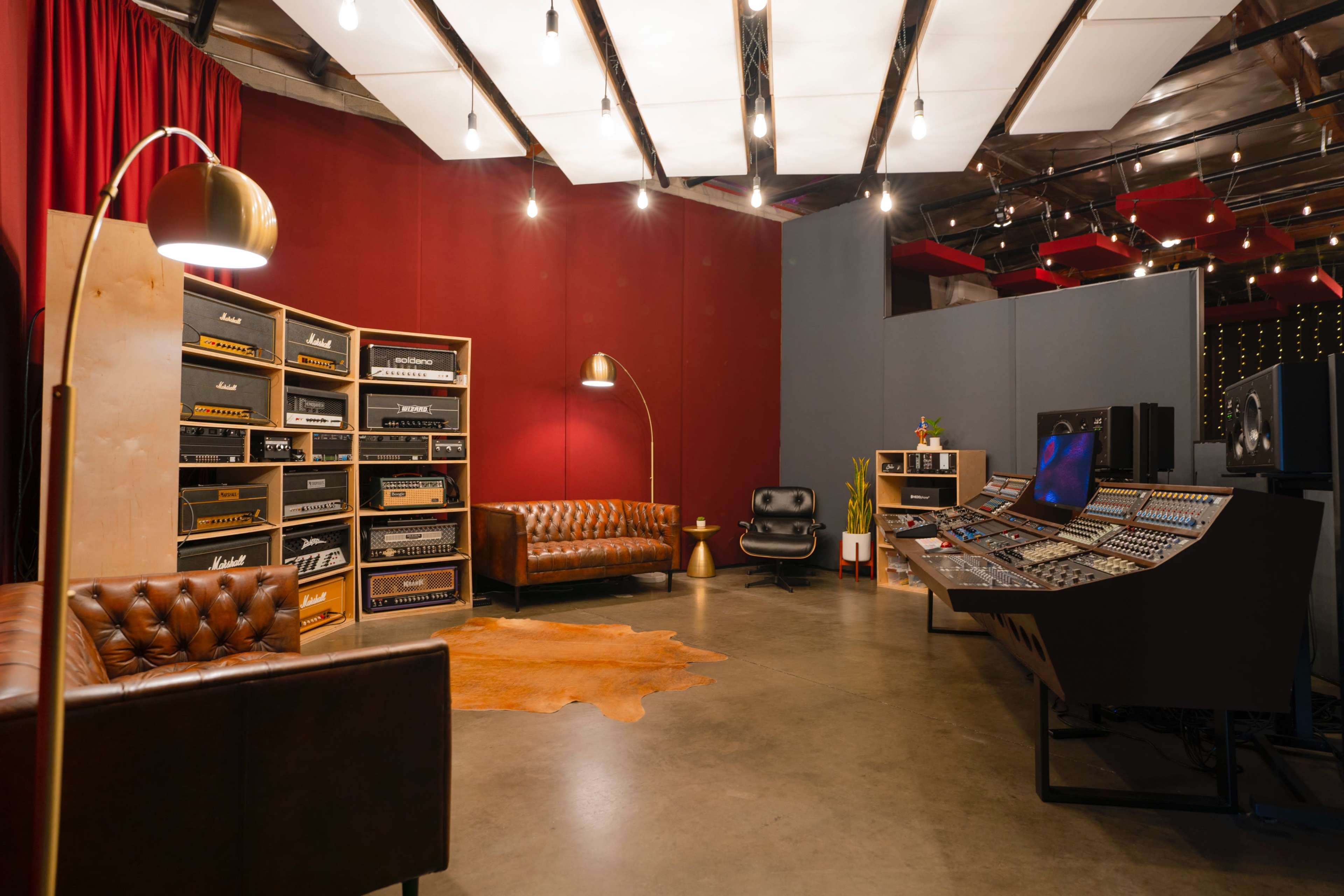 A modern recording studio featuring a brown leather couch, various amplifiers against a red wall, and a sound mixing console with overhead lighting.
