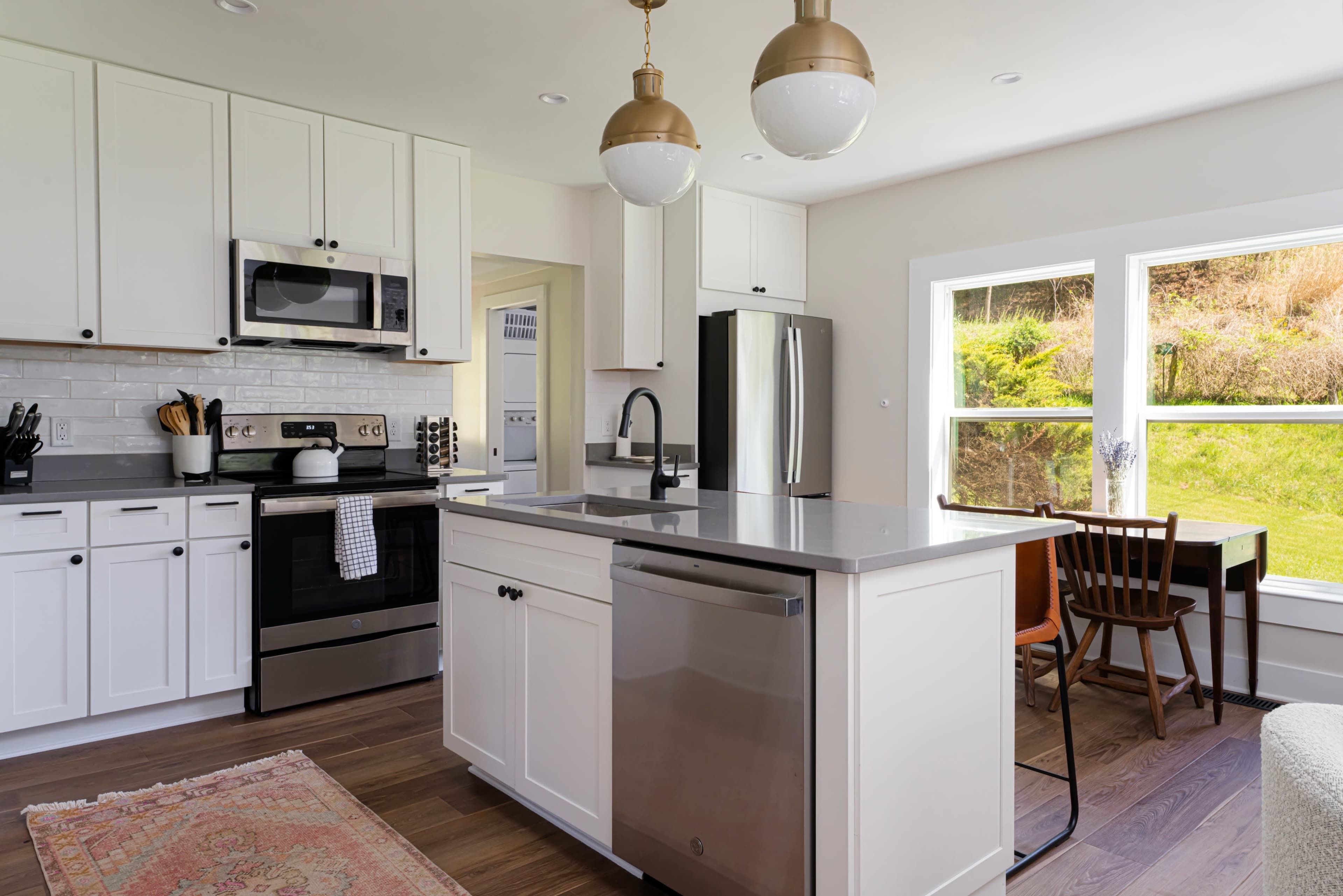 The image shows a modern kitchen with white cabinetry, stainless steel appliances, a central island with a gray countertop, and a window overlooking a green outdoor landscape.