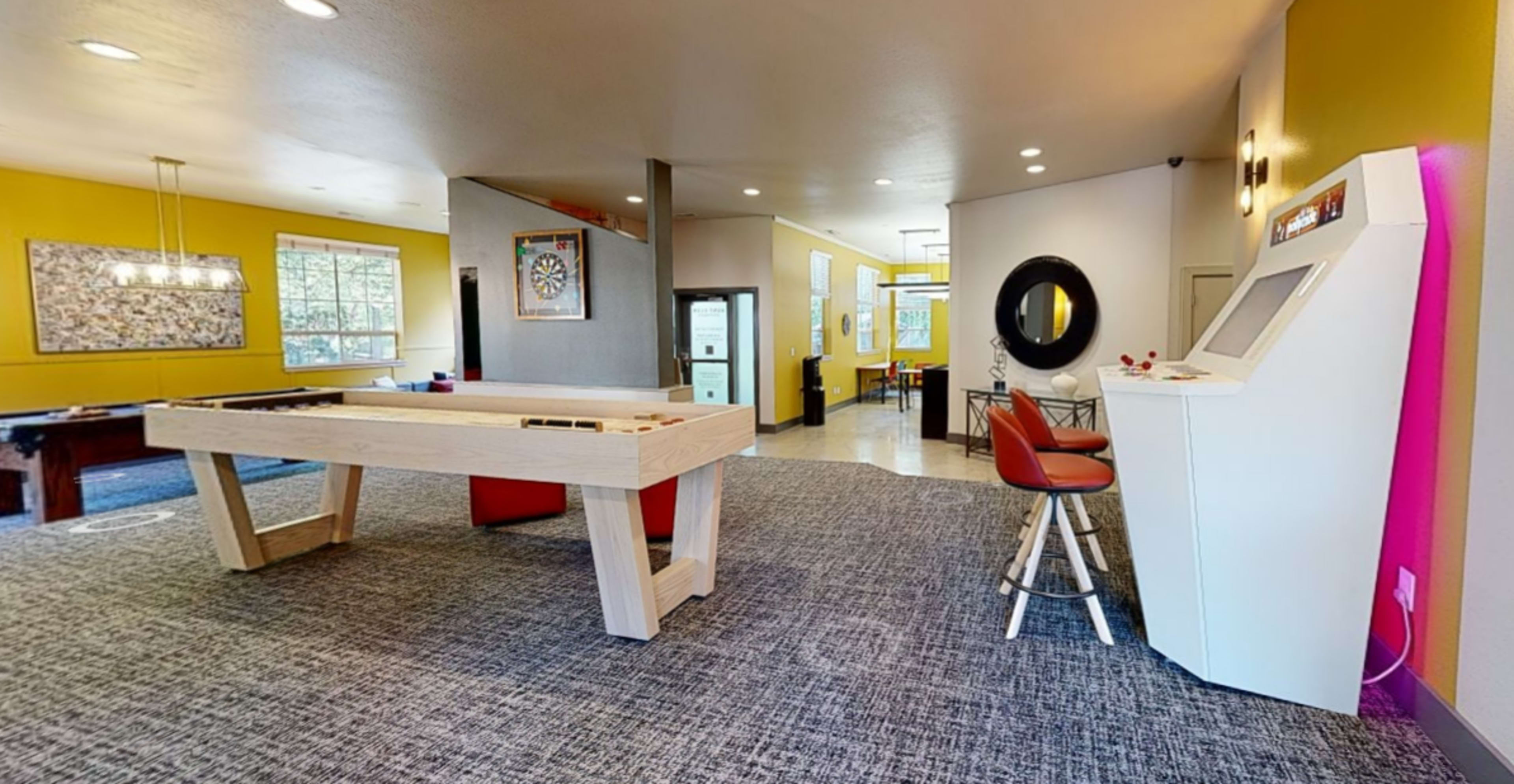 A spacious room featuring a shuffleboard table, a seating area with a bar stool, and a digital game console against a backdrop of yellow walls and artwork.