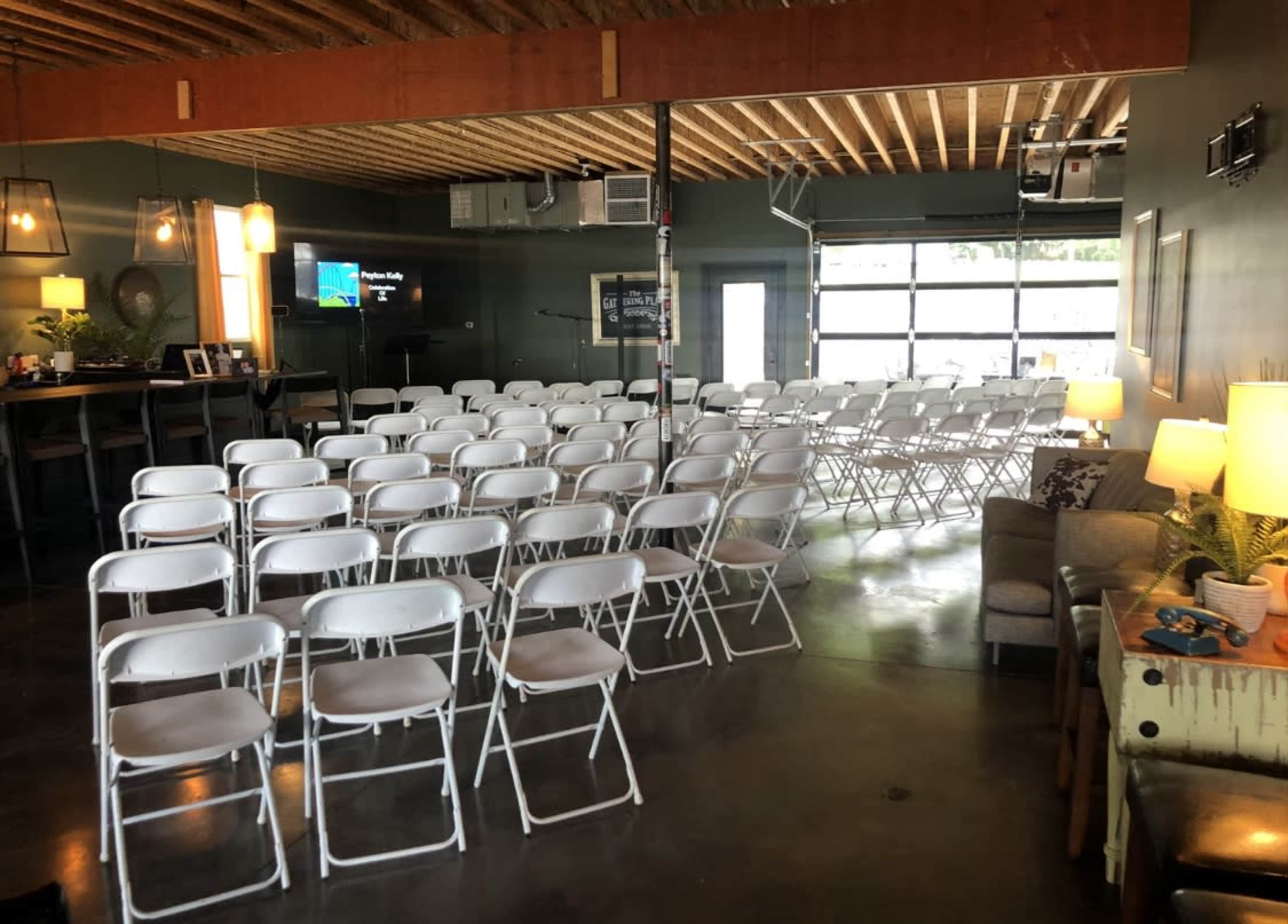 The image shows a room set up with rows of white folding chairs facing a screen, with a casual seating area and a bar in the background.