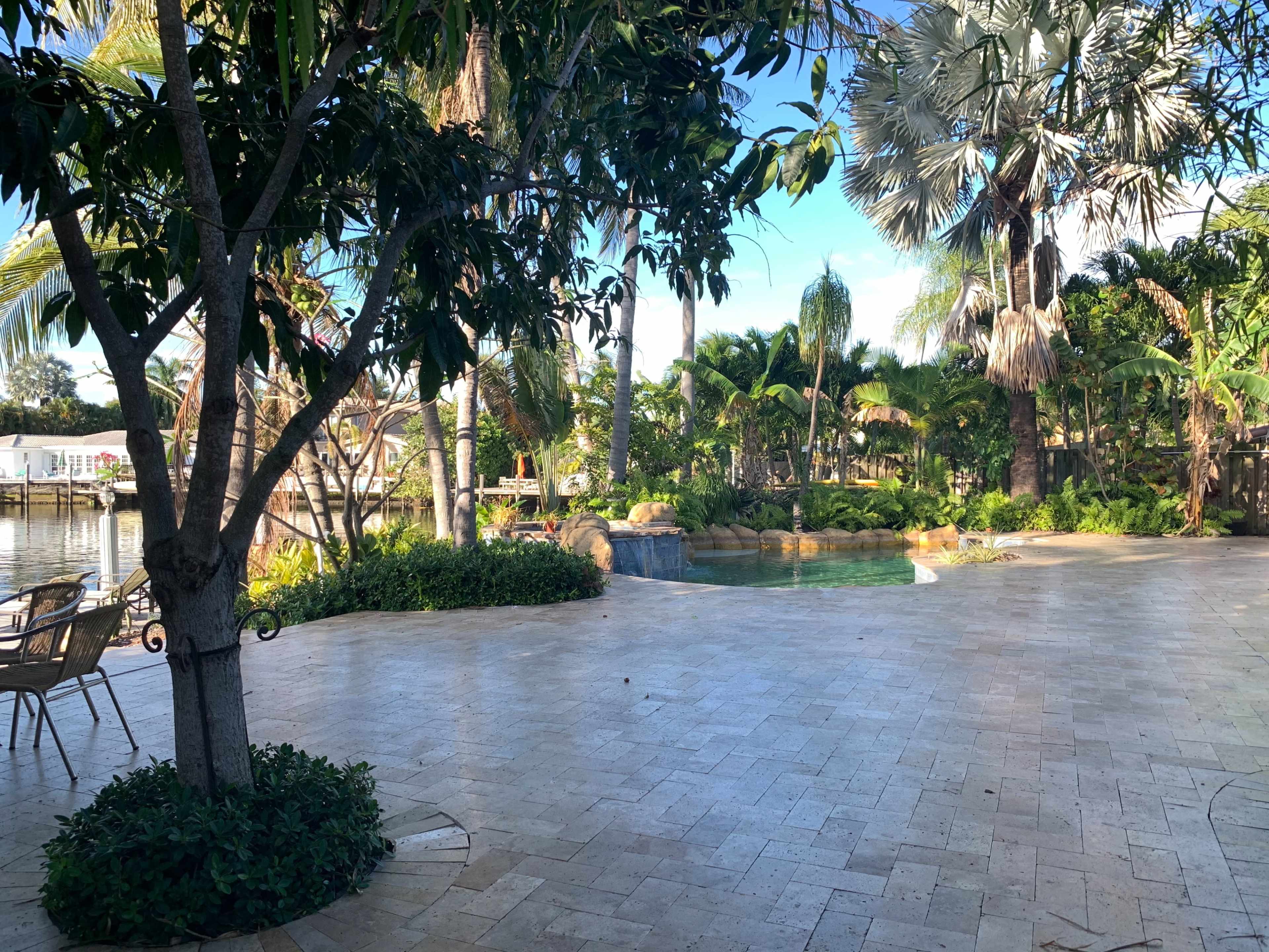 The image shows a spacious patio area surrounded by tropical plants and a swimming pool with a water feature.