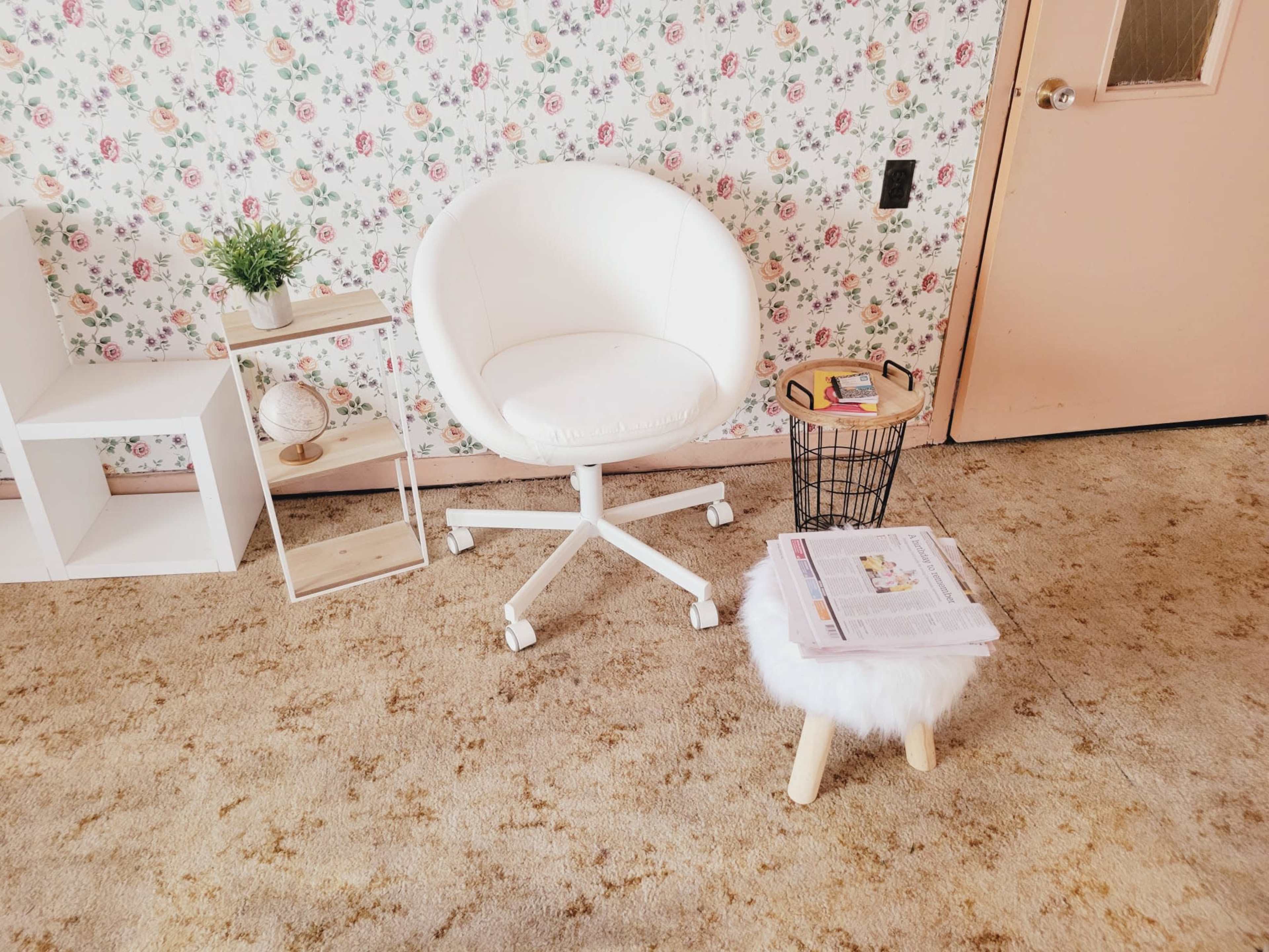 The image shows a small living space with a white swivel chair, a round stool with a fluffy seat, a small side table holding a magazine, and a decorative shelf against a floral wallpaper background.