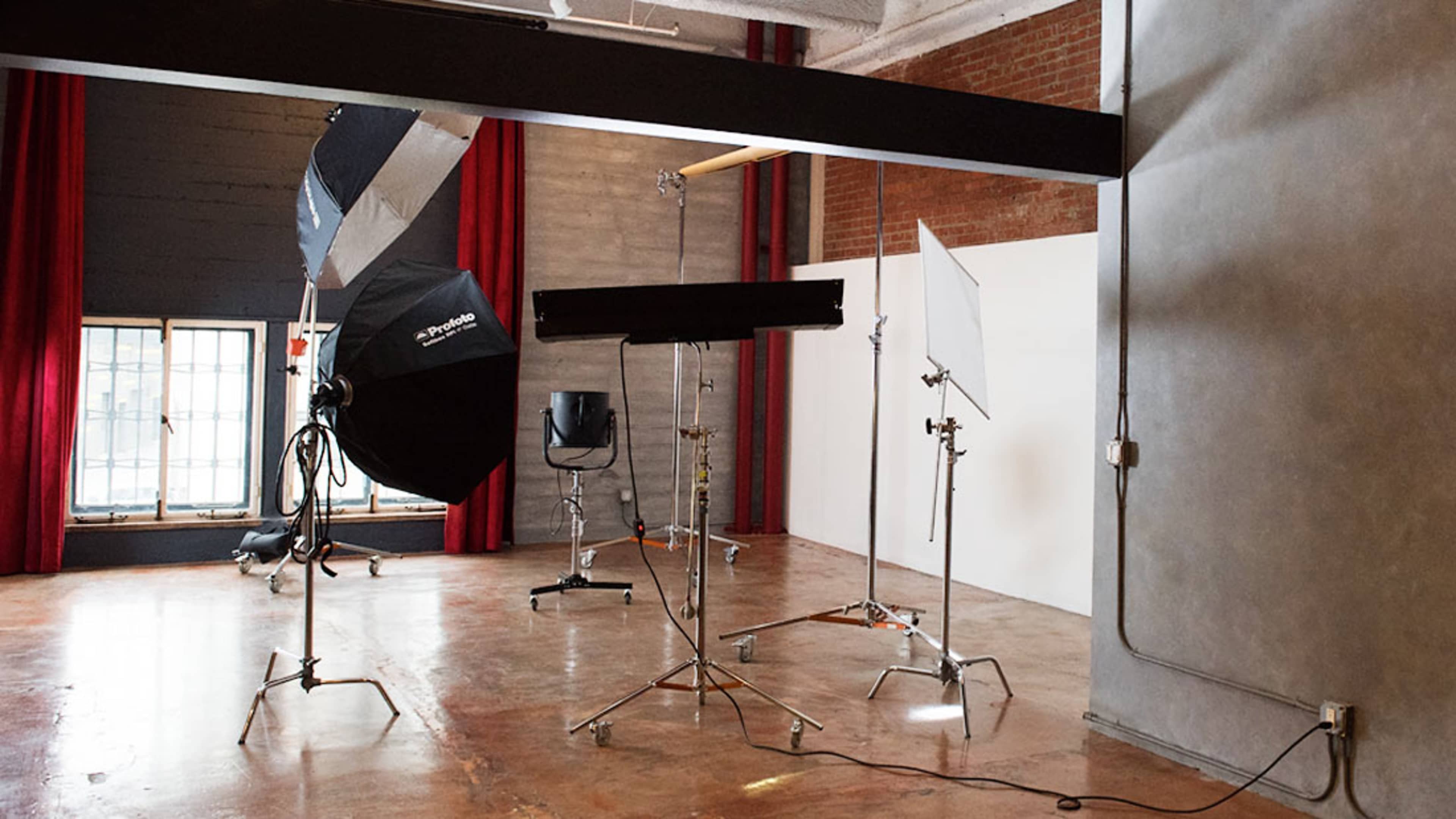The image shows a photography studio with umbrellas, softboxes, and a backdrop setup, featuring a chair and large windows with red curtains.