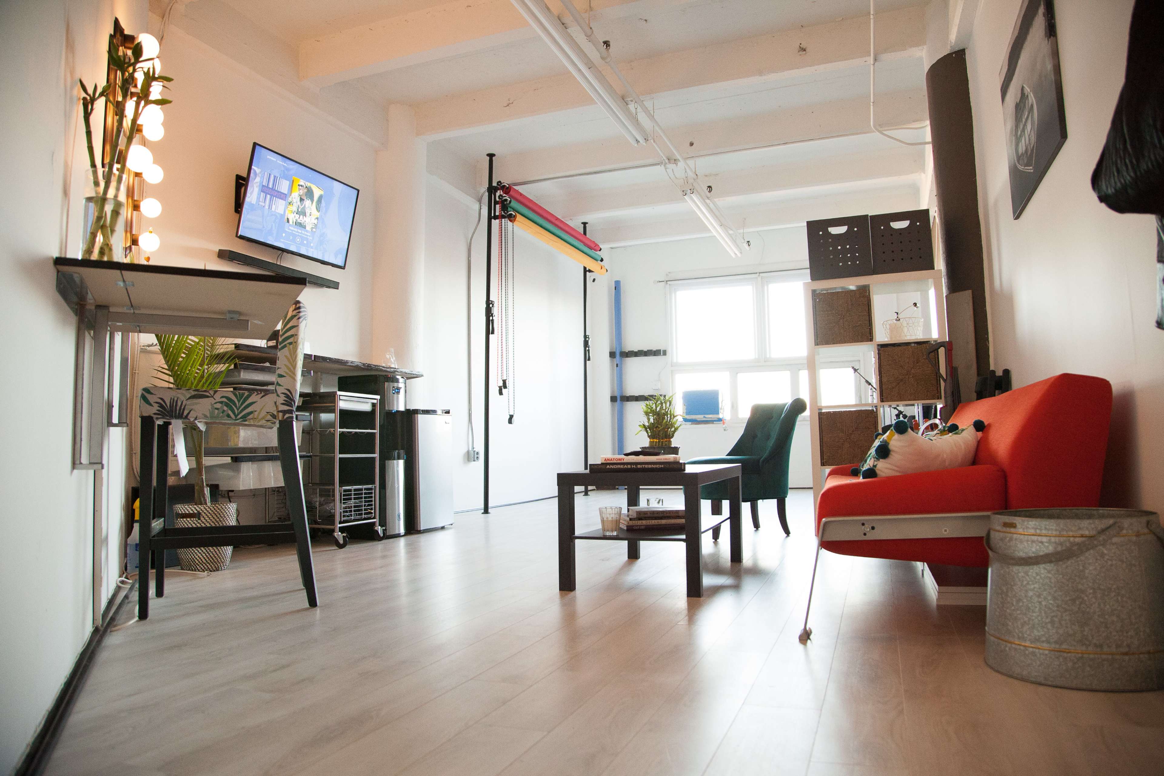 The image shows a modern, minimalistic living space featuring a red sofa, a coffee table, a small dining area, and a workspace with a TV mounted on the wall.