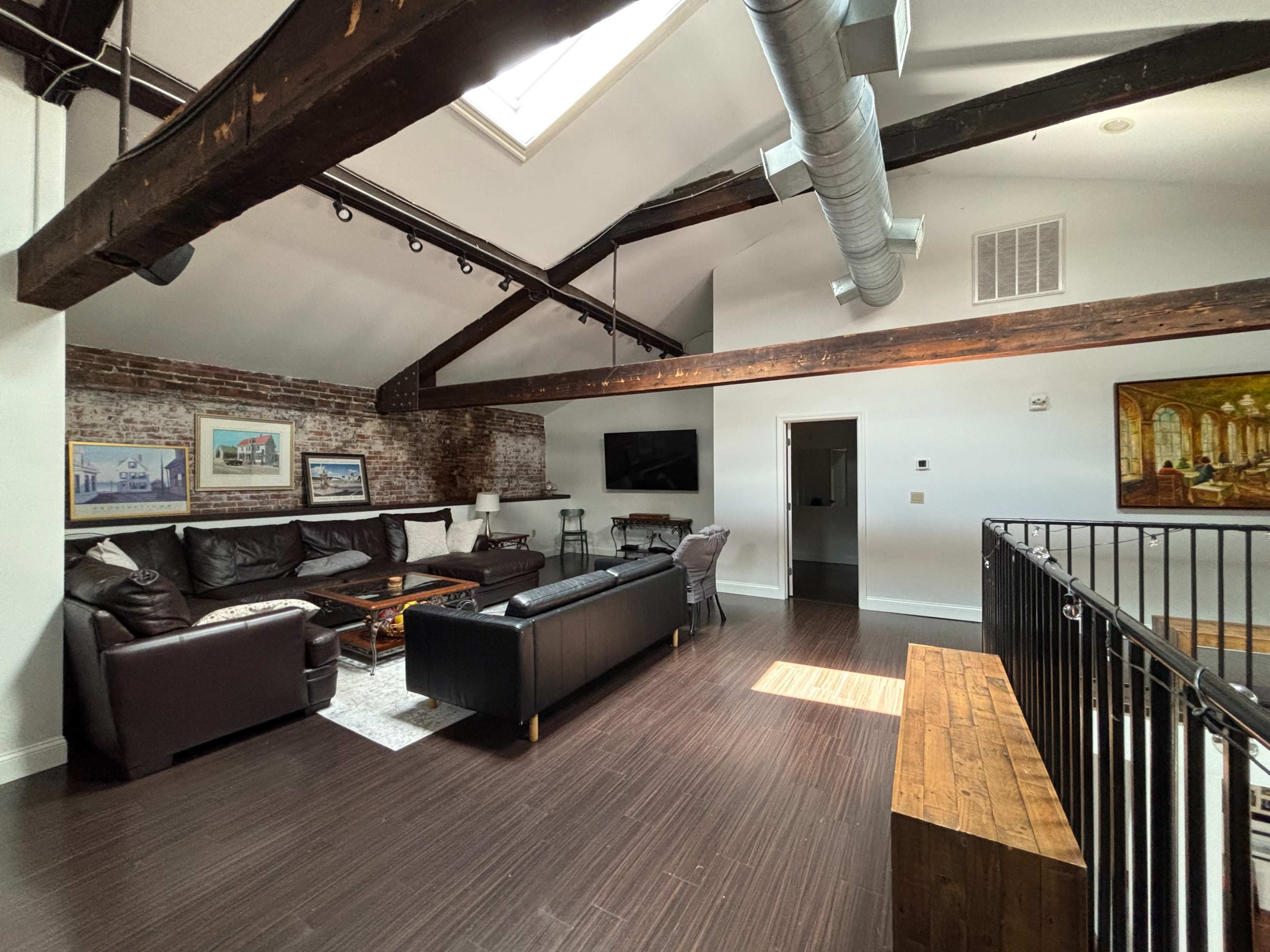 The image shows a spacious living area with exposed brick walls, wooden beams, and modern furnishings, including a sectional sofa and large windows.