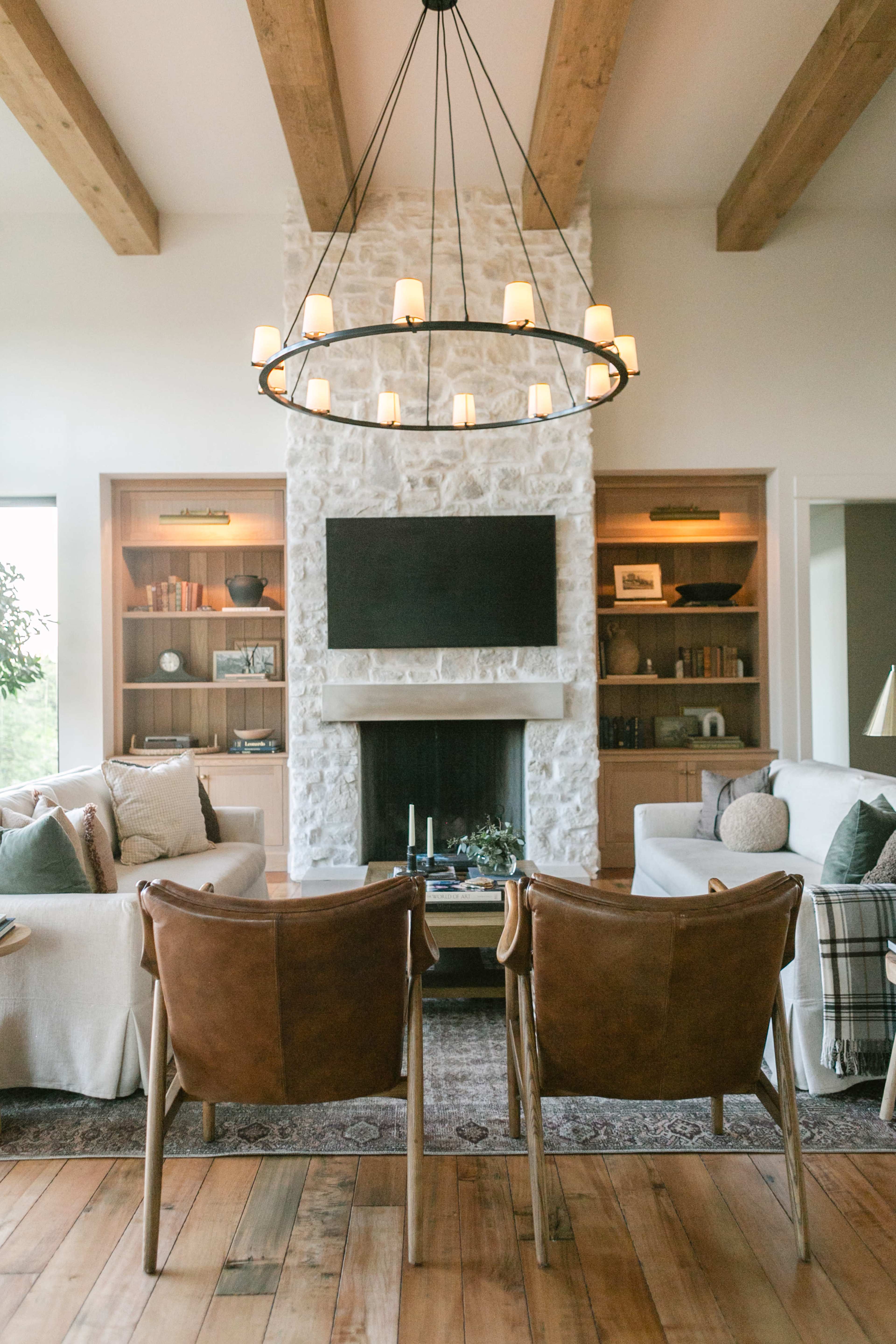 The image shows a cozy living room with a stone fireplace, a large circular chandelier, and two leather chairs facing a television.